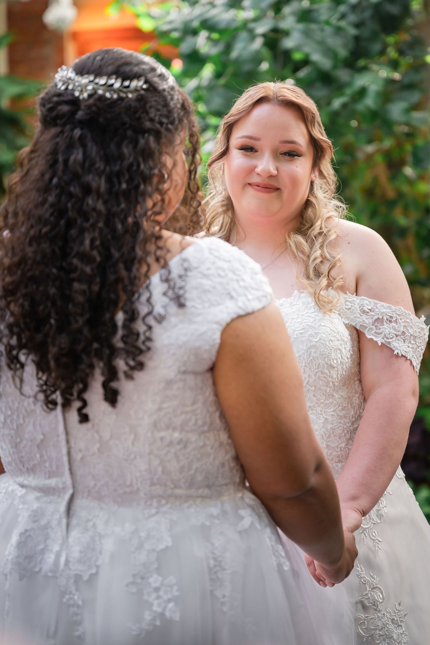 Two beautiful brides in white lace wedding dresses share an intimate moment in a garden setting.