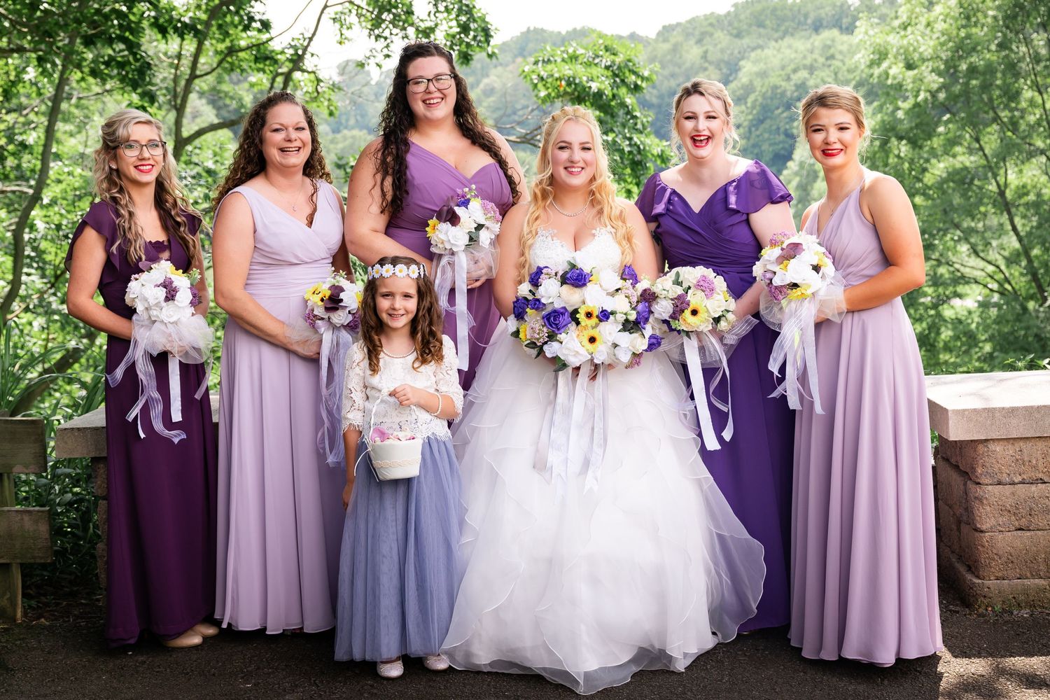 Wedding party in purple dresses poses together with bouquets on stone terrace.