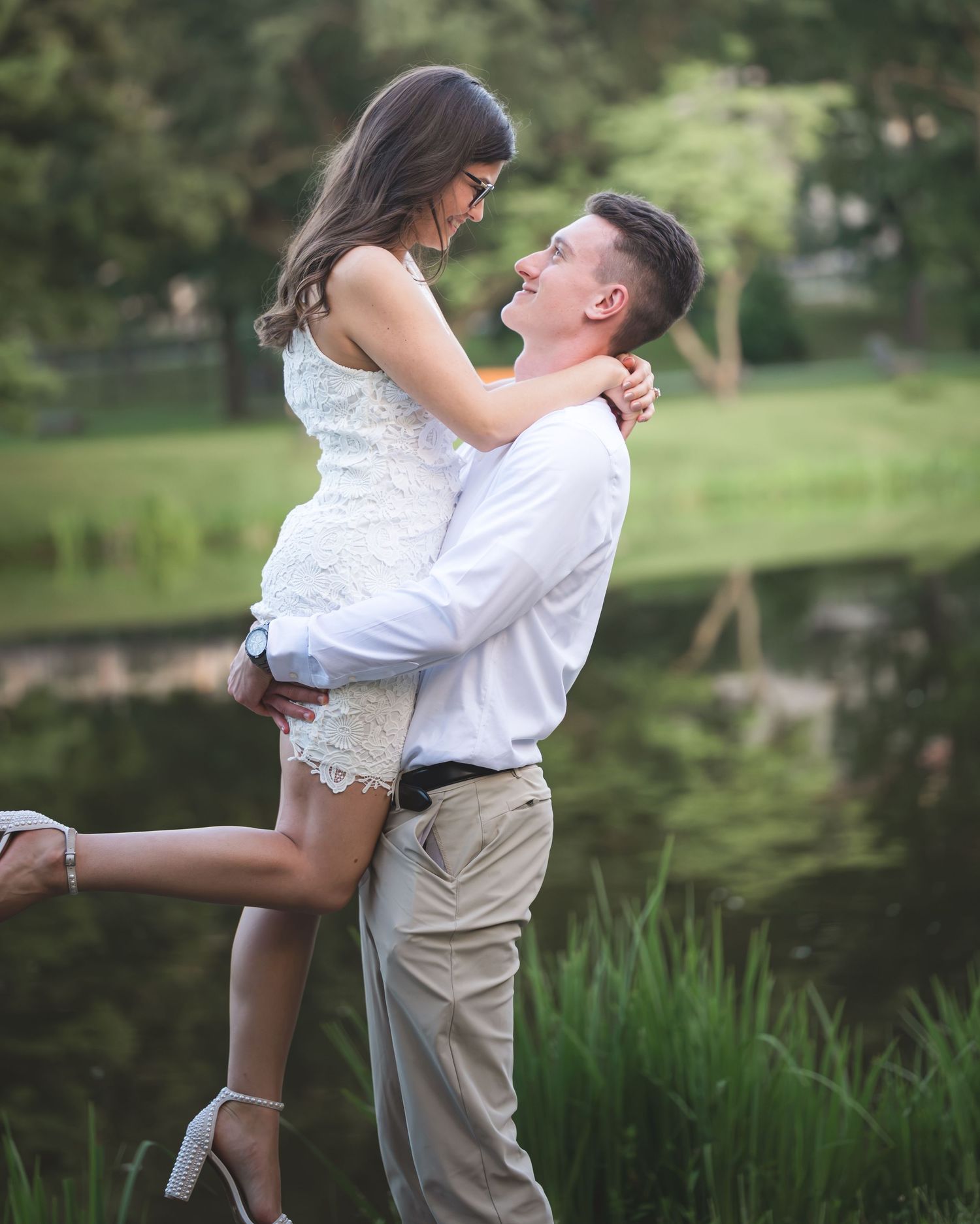 A couple shares a romantic moment outdoors, man lifting woman in white dress near a serene pond surrounded by lush greenery.