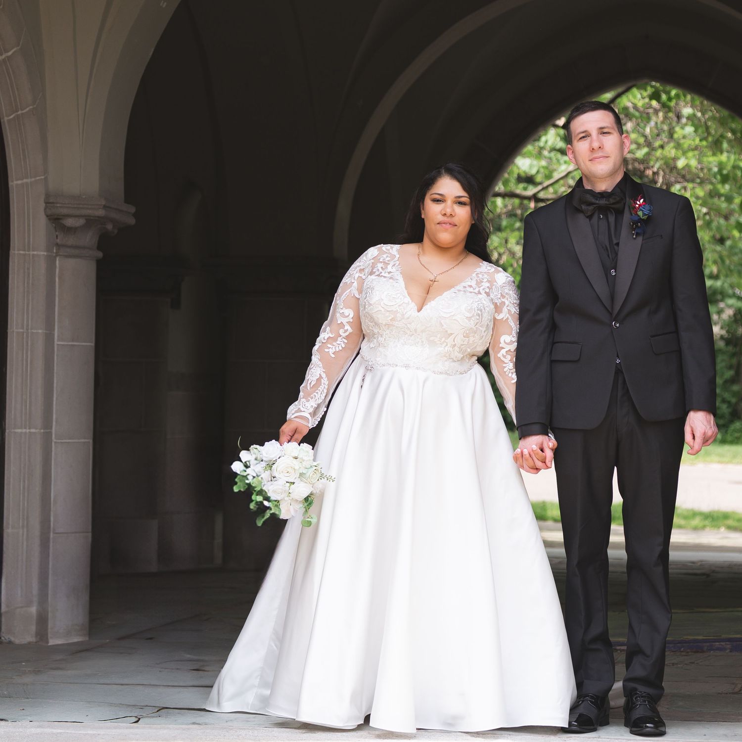 A couple poses under stone archways during their wedding, with the bride in a lace sleeve gown holding white flowers.