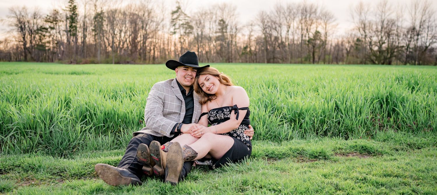 A couple sits together on a blanket in a lush green field during golden hour at sunset.