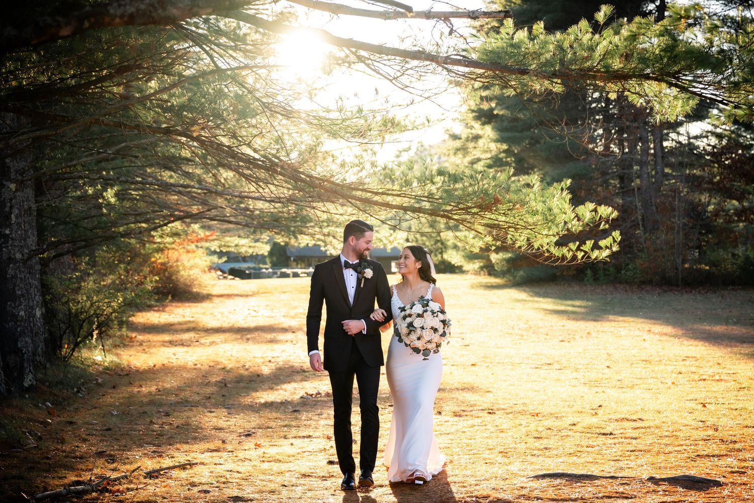 A couple in wedding attire walk down a sunlit forest path during golden hour.