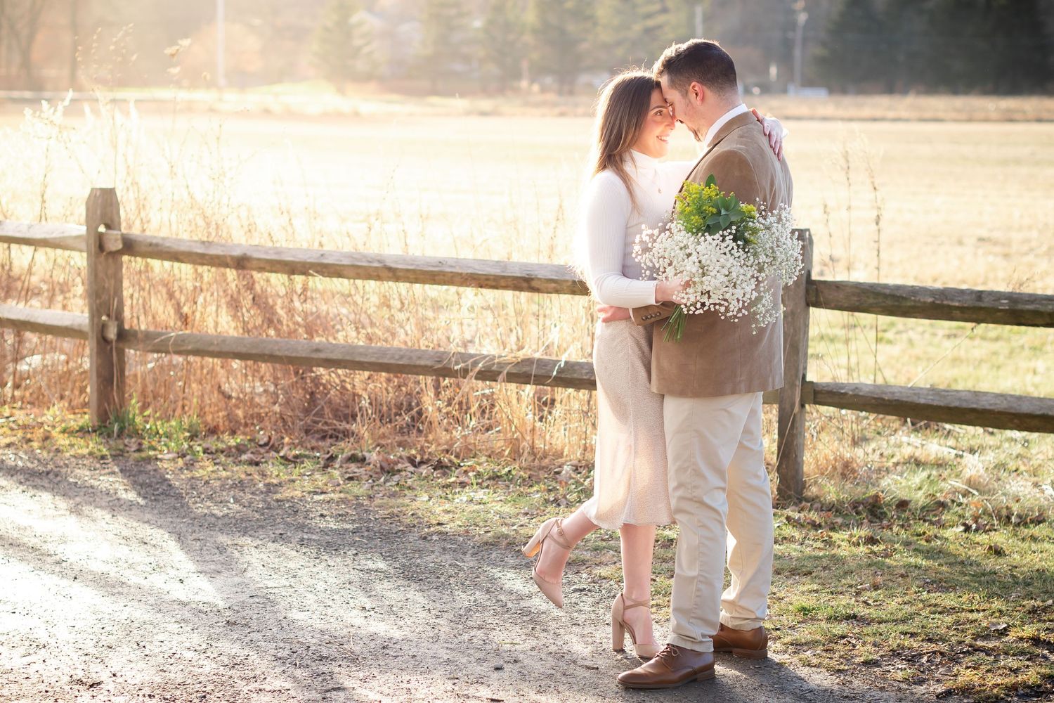 A couple shares an intimate moment on a sunlit path near a rustic wooden fence.