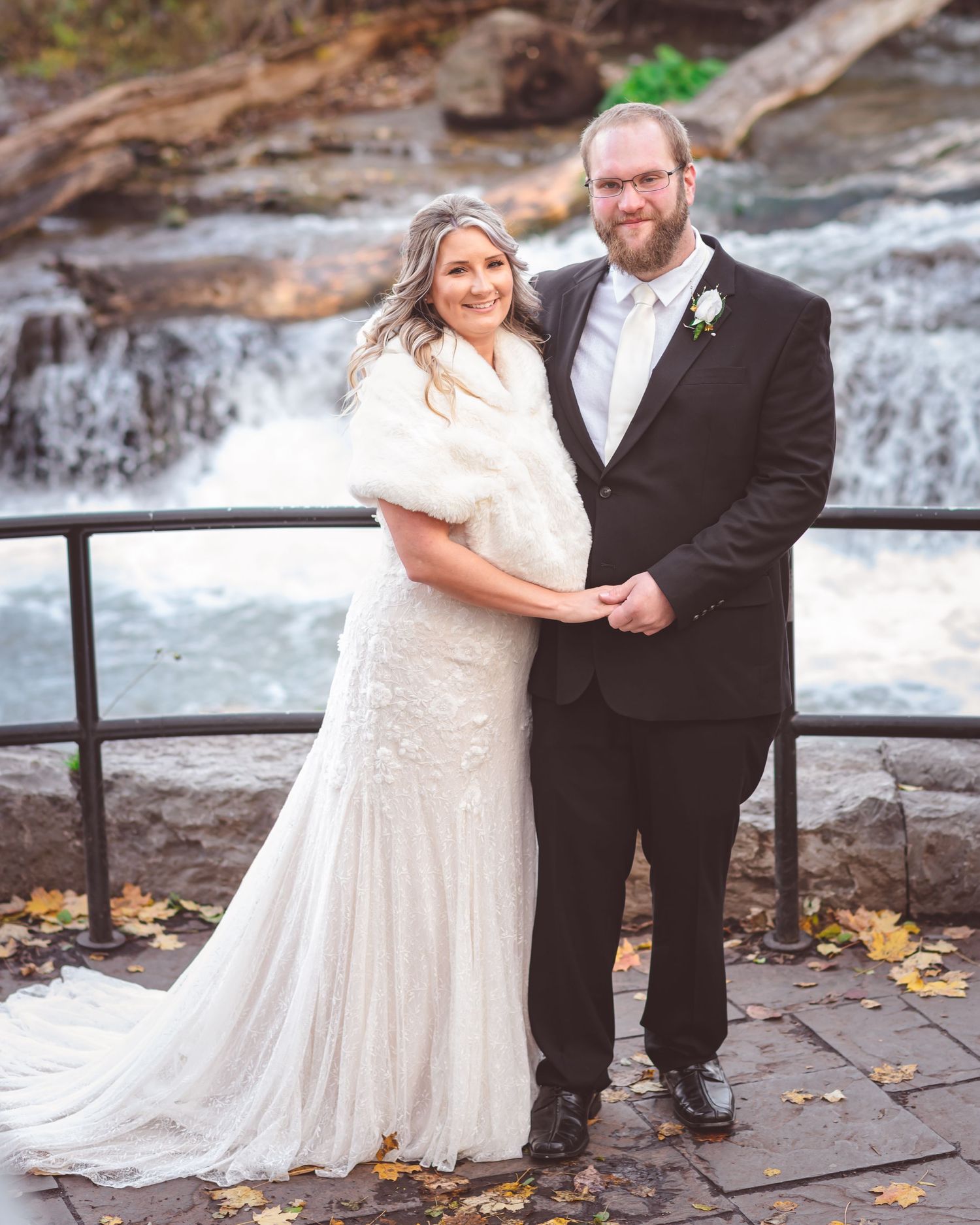 Wedding couple poses together on viewing platform with waterfall and autumn scenery in background.