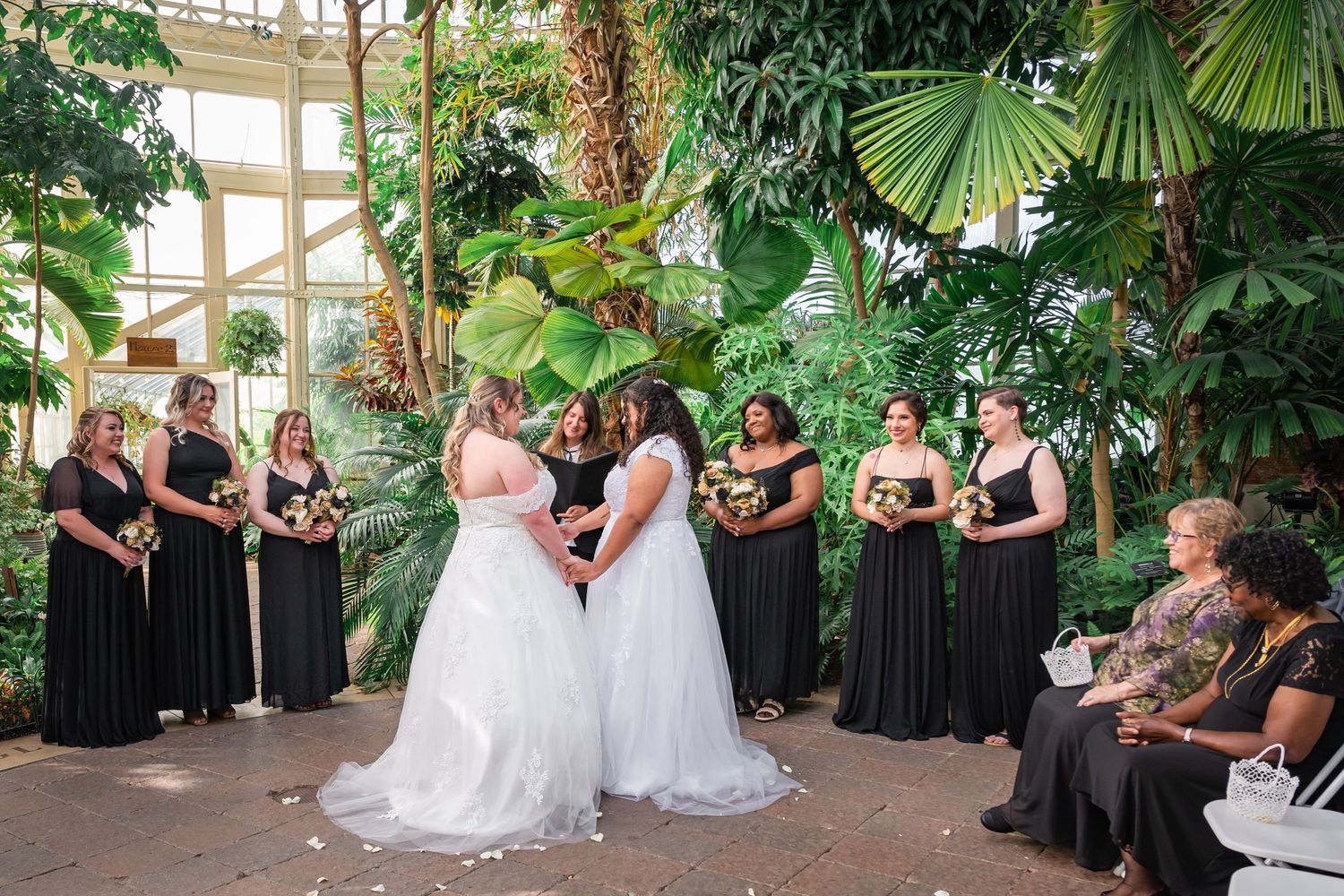 Wedding ceremony in a botanical garden with attendants in black dresses surrounded by lush greenery.
