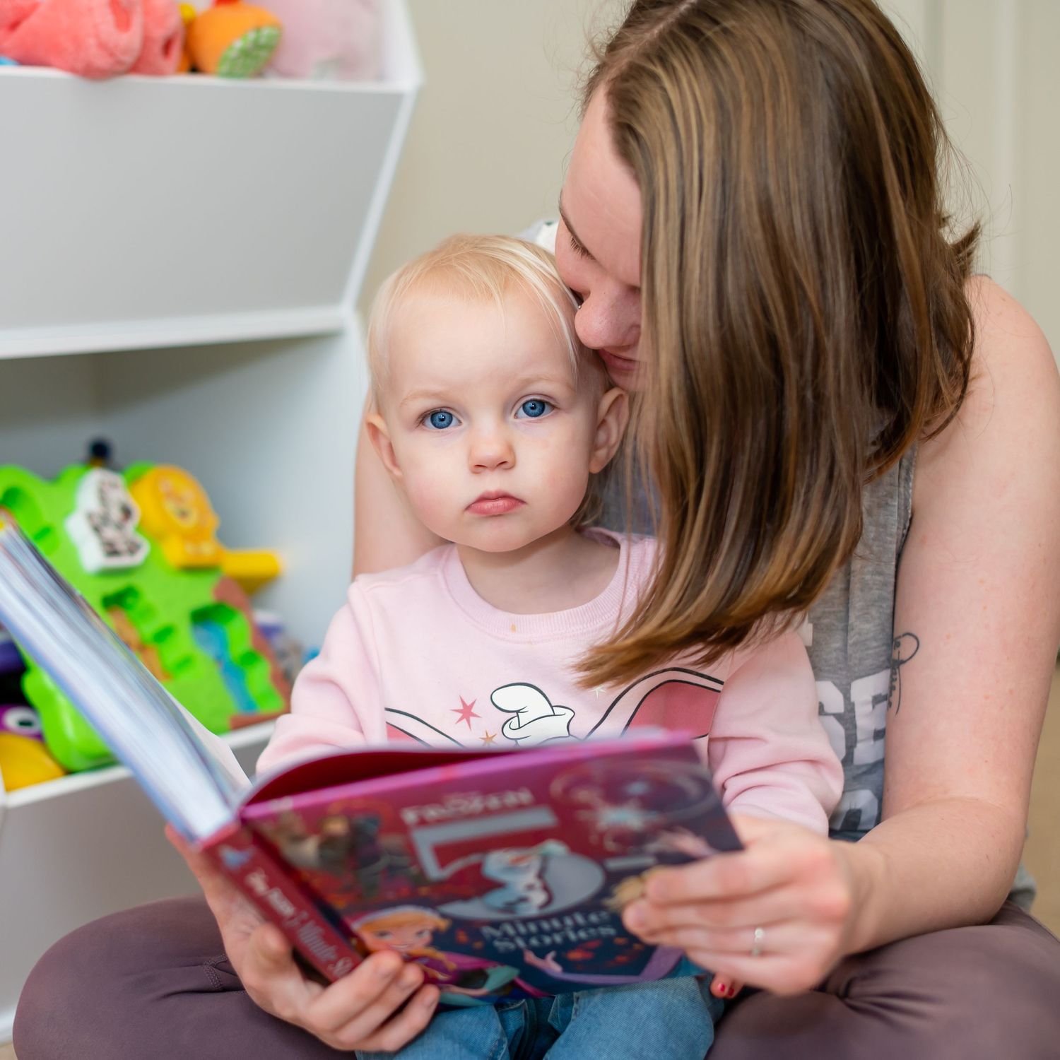 Parent and child reading a book together in playroom with toy shelves.