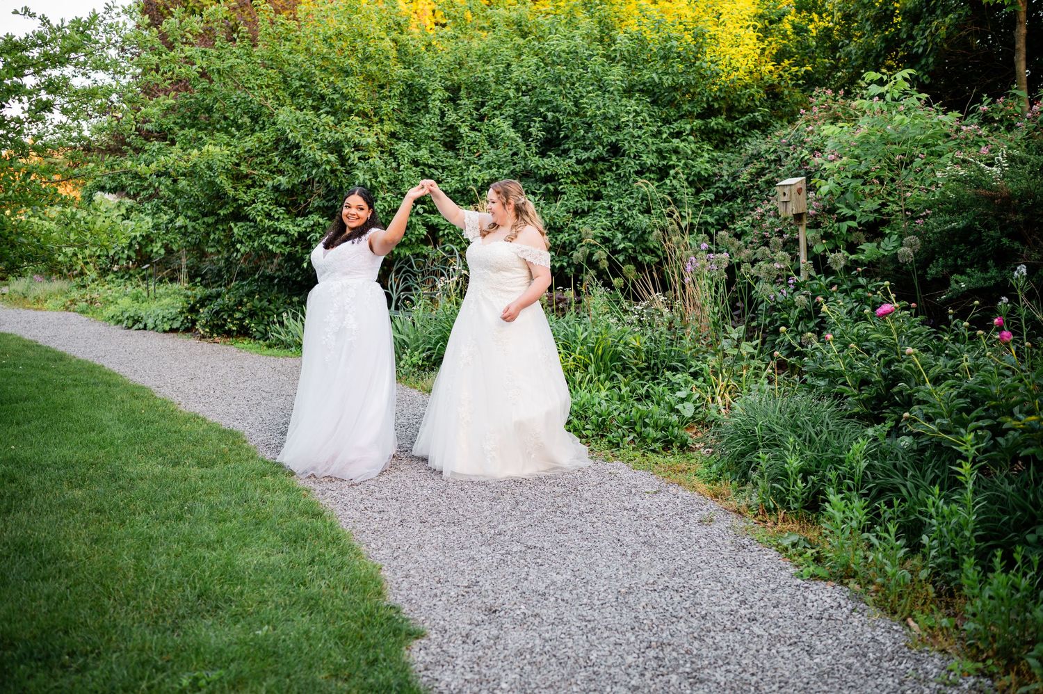 Two brides in flowing white dresses pose together on a garden path with yellow flowers in background.