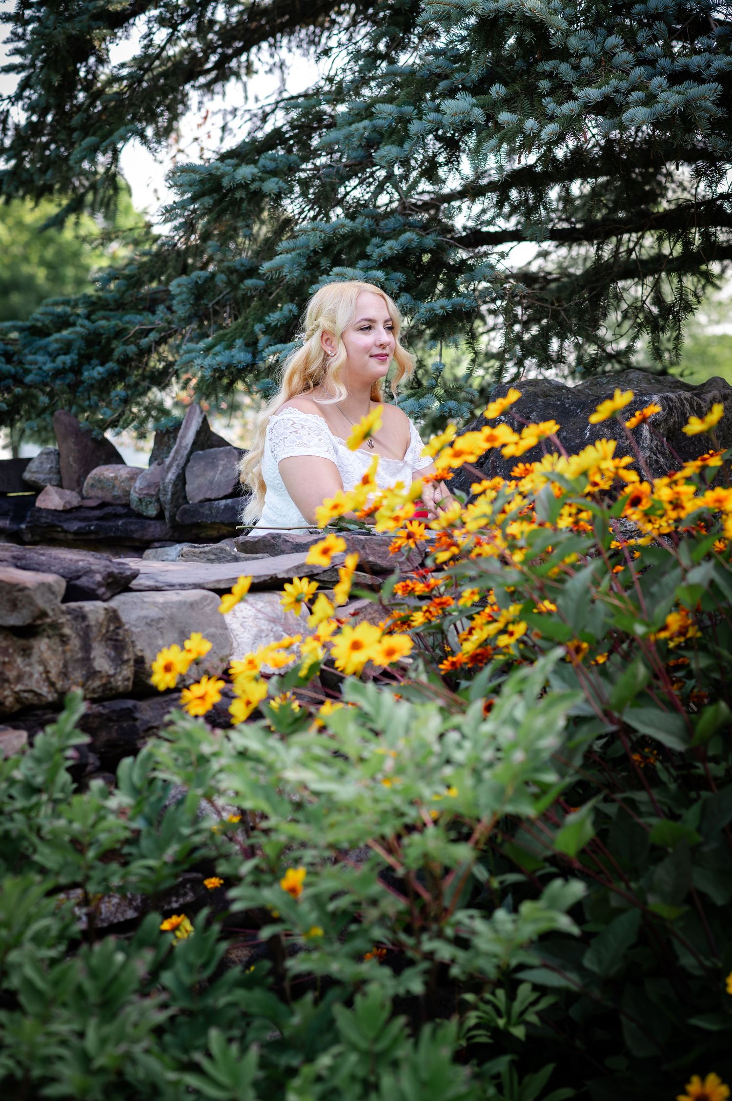 Figure in white sits by stone wall with yellow flowers and evergreen trees in background.