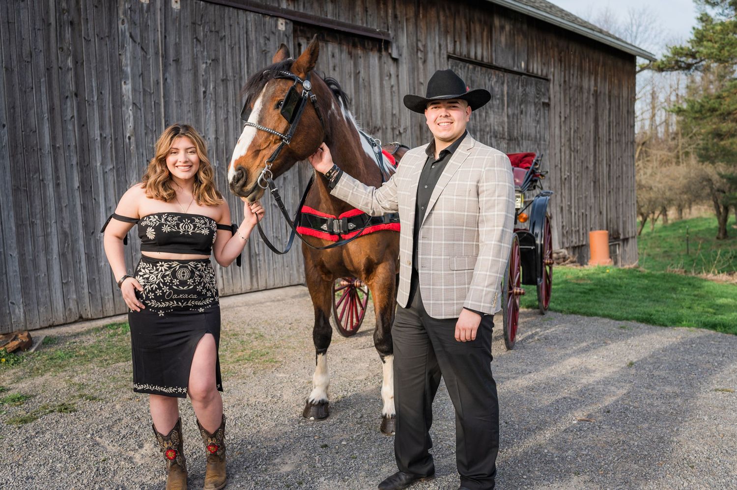 Western-styled couple poses with saddled horse outside rustic barn.
