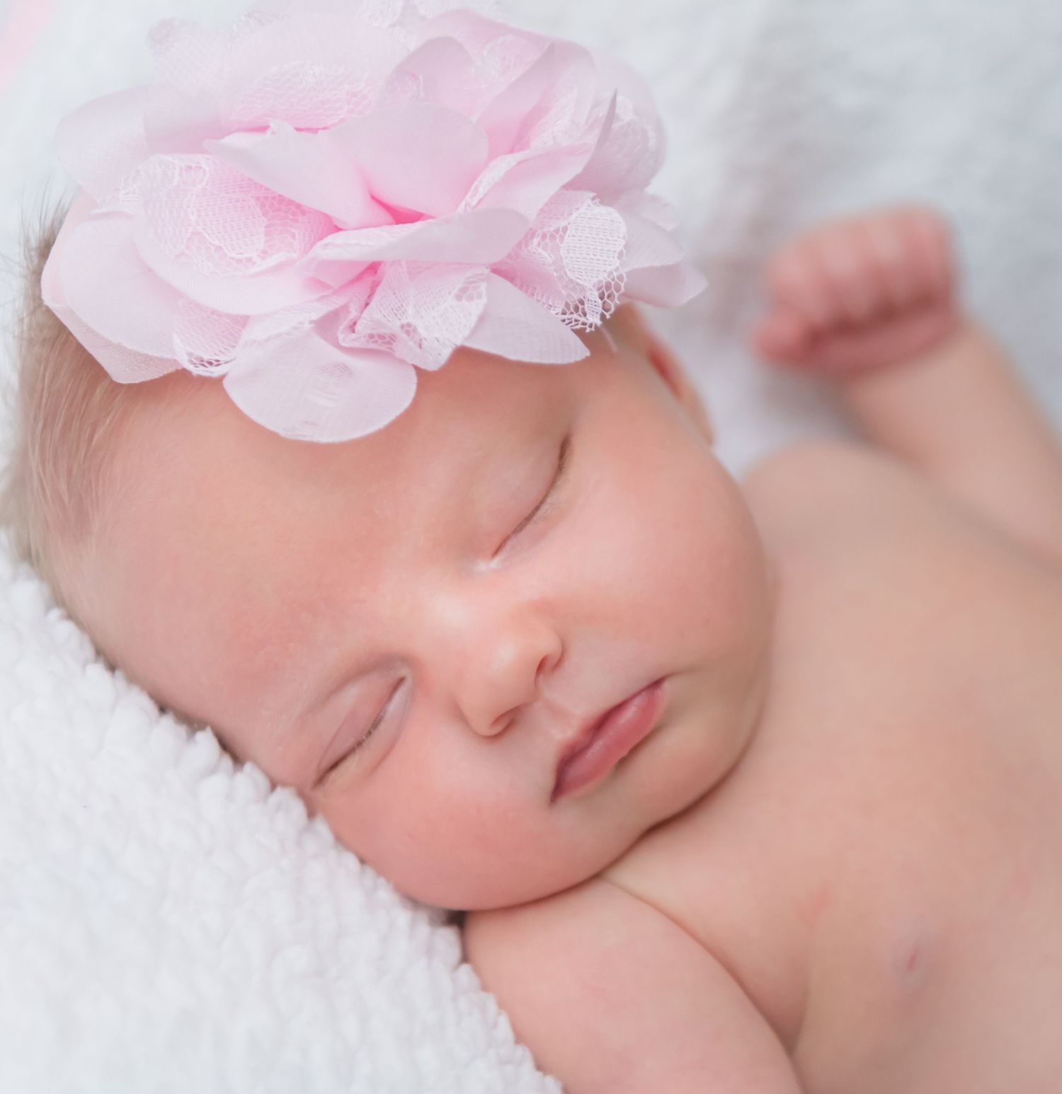 Sleeping newborn baby wearing pink floral headband rests peacefully on white blanket.