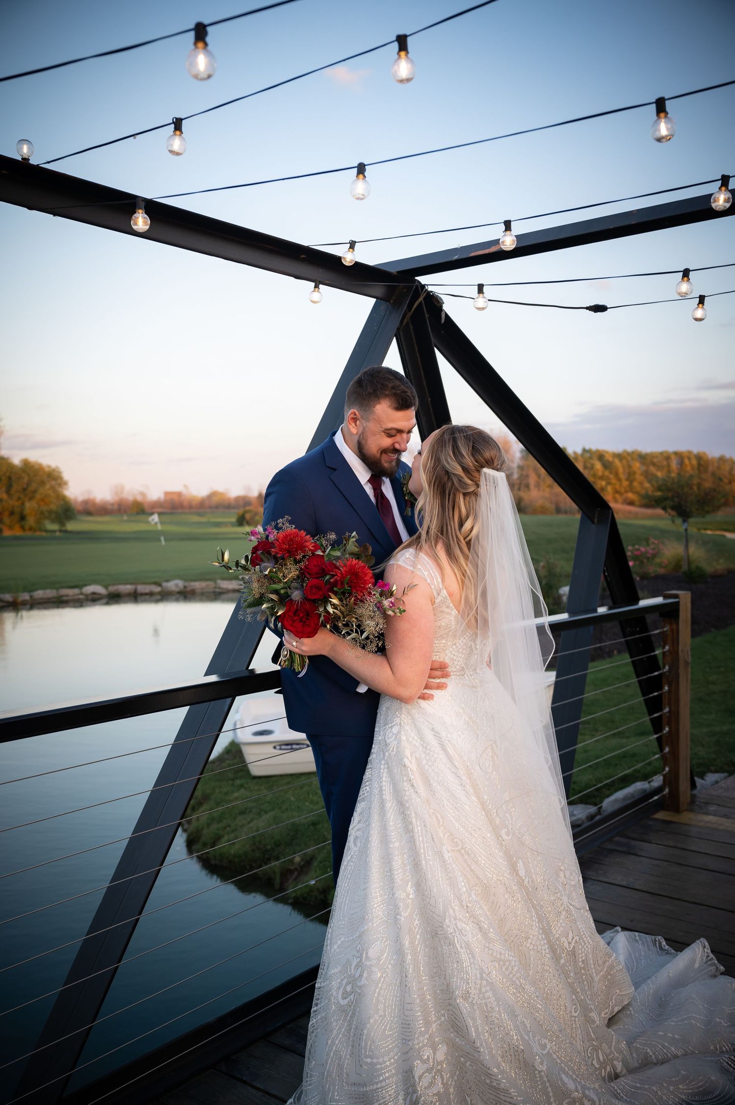 Newlyweds share intimate moment on wooden deck with string lights and lake view at dusk.
