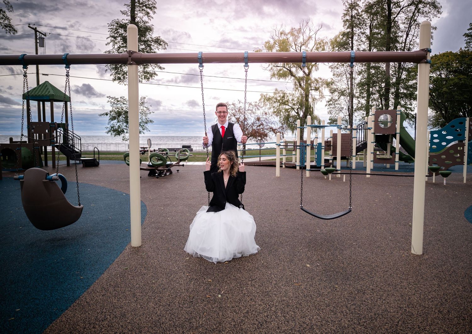 Playful wedding photo at playground with couple on swings near lakefront.