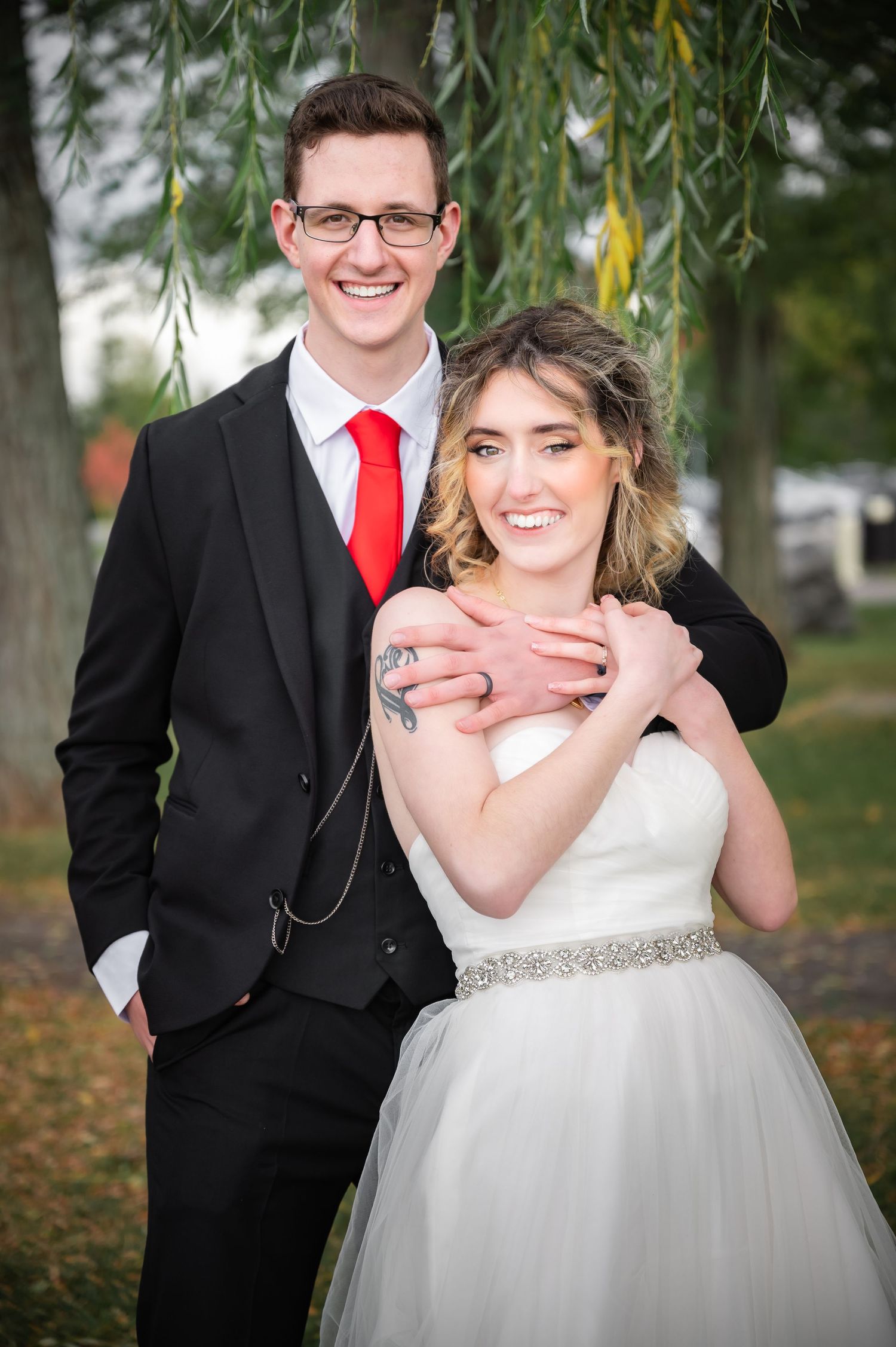 A joyful wedding couple poses under a willow tree, with the groom in black suit and red tie.