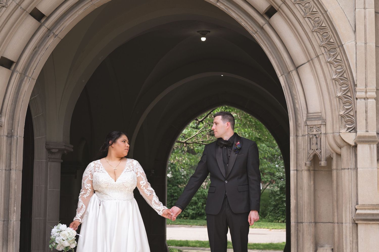 A wedding party underneath stone archways in an elegant outdoor setting, surrounded by greenery and architectural details.