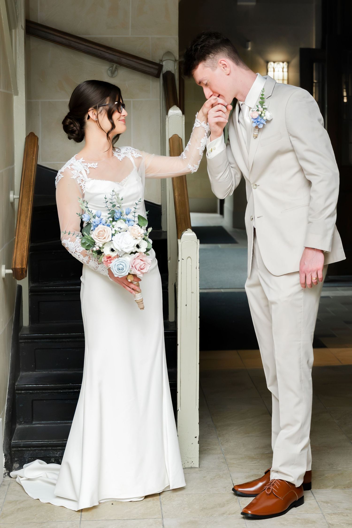 Groom in beige suit kisses bride's hand as she holds pastel bouquet on wedding day near staircase.