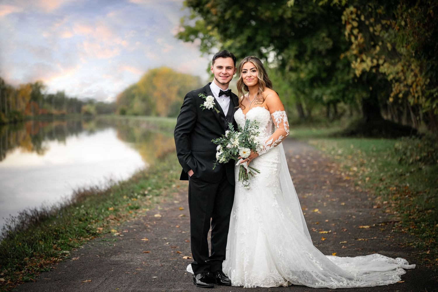 A couple in formal wedding attire poses together on a scenic autumn path by a tranquil river.