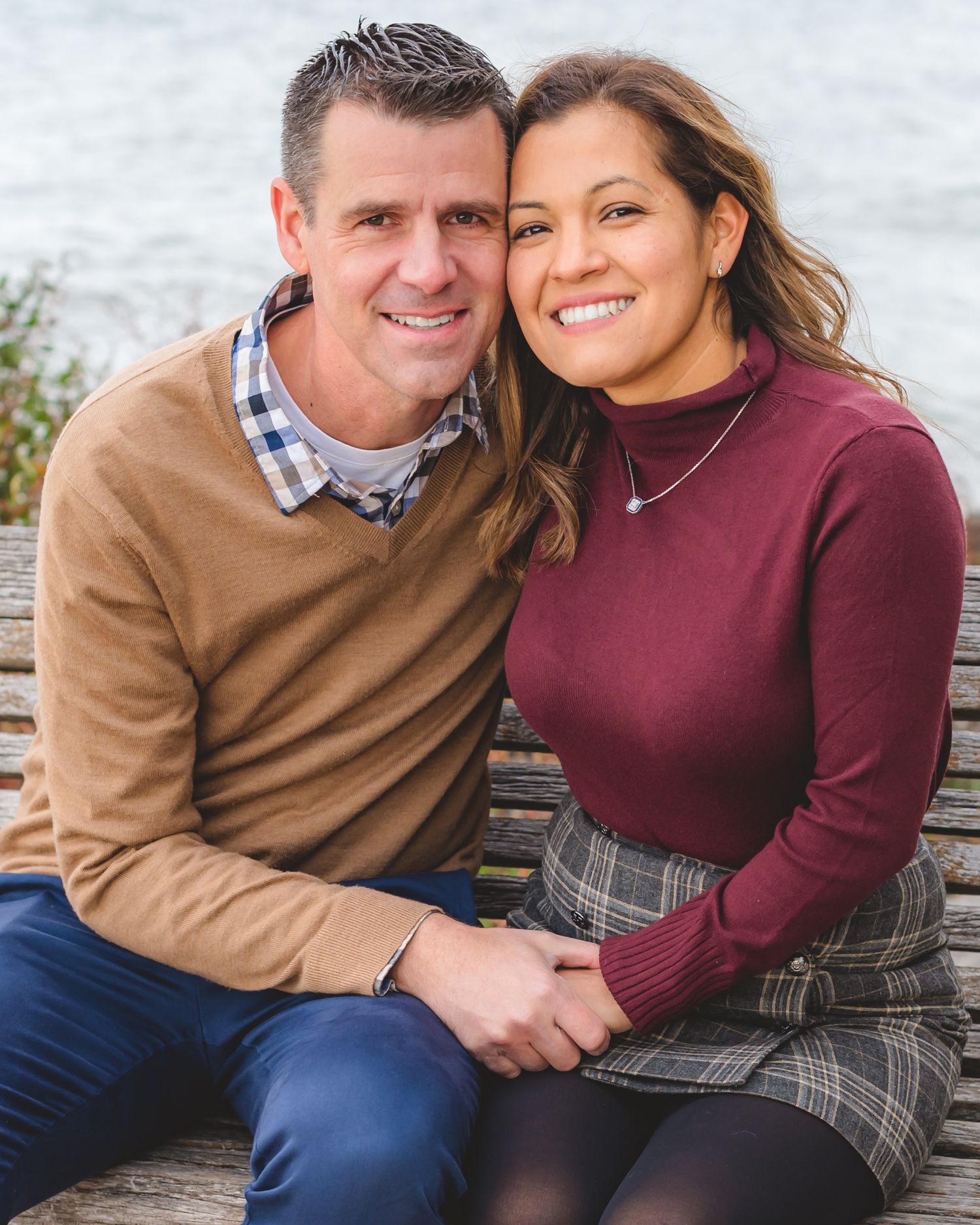 A couple wearing burgundy and tan sweaters smile together while sitting outdoors on a bench.