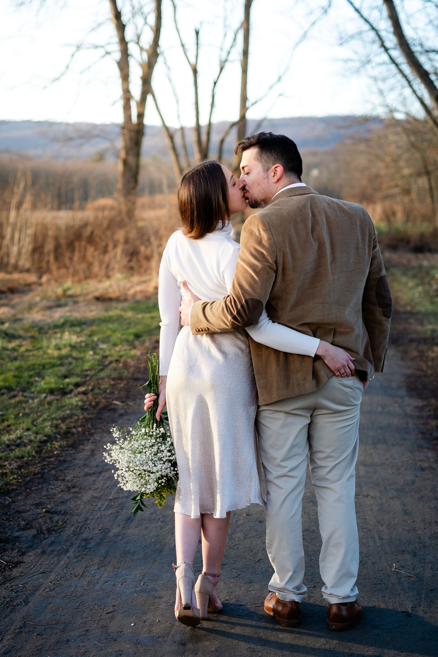 Couple walking along a scenic mountain trail in warm evening light holding a delicate bouquet.