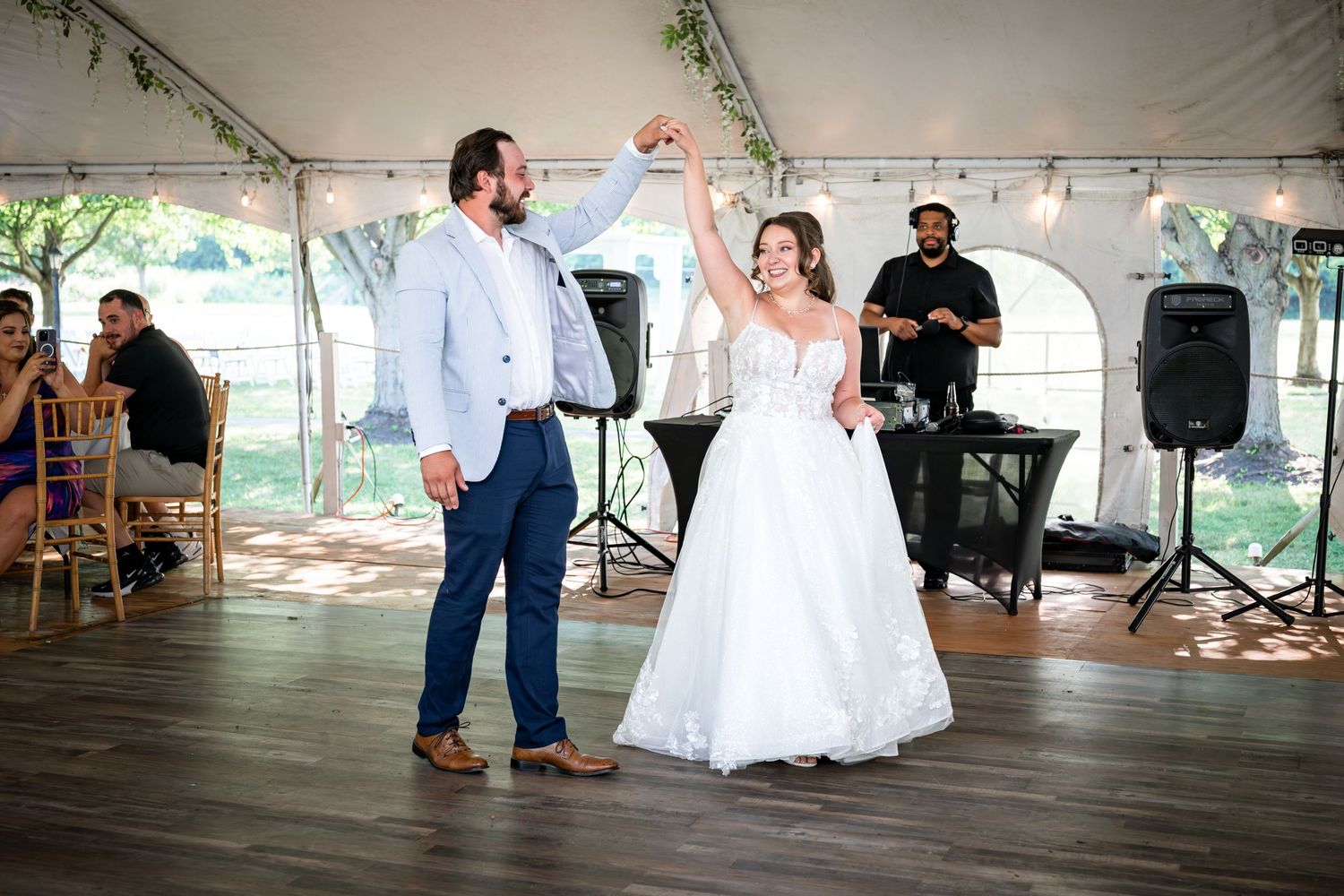 Bride and groom share first dance under white tent at outdoor wedding reception with DJ in background.