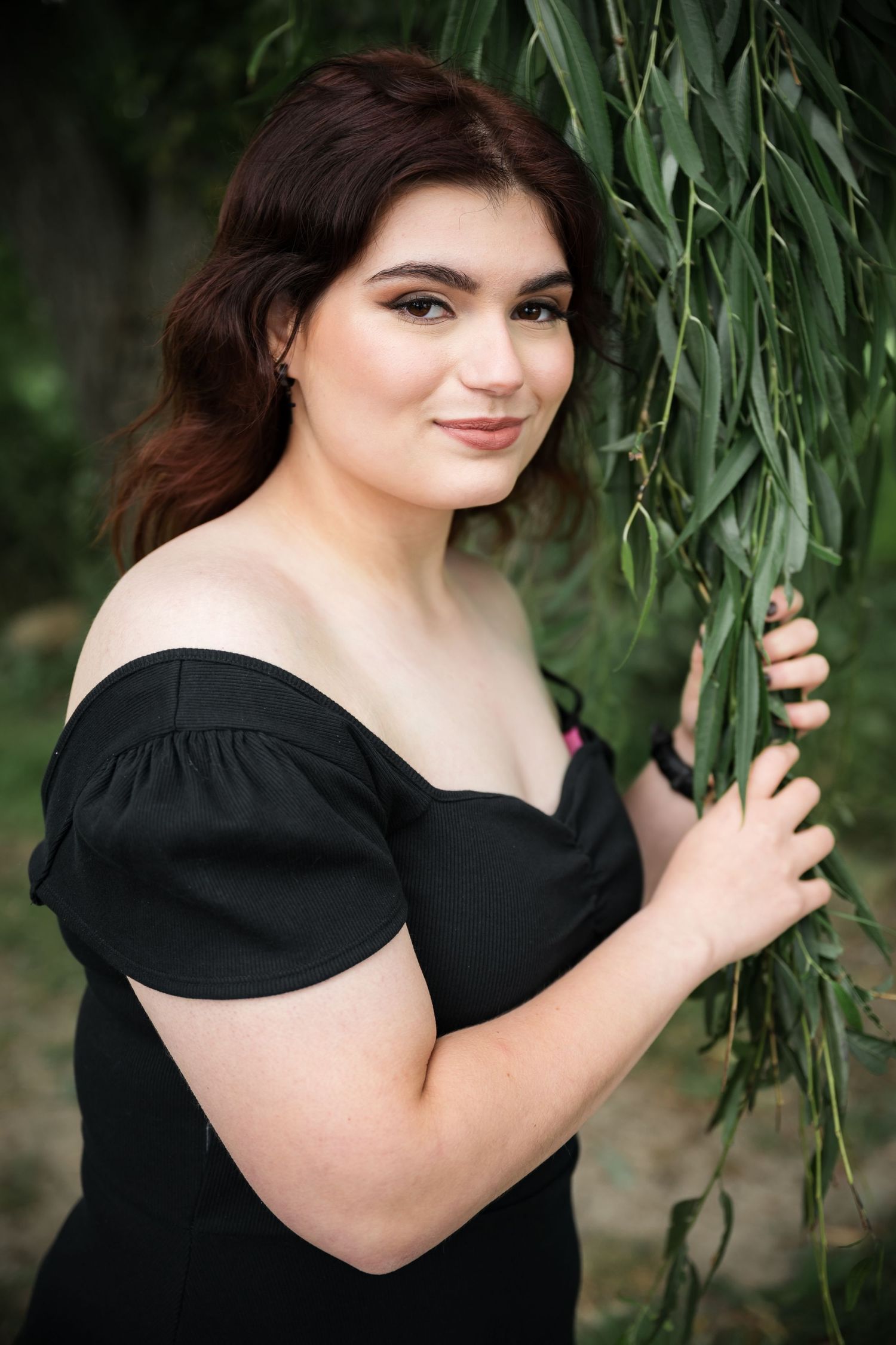 Woman in black off-shoulder dress poses elegantly by willow branches in a garden setting.