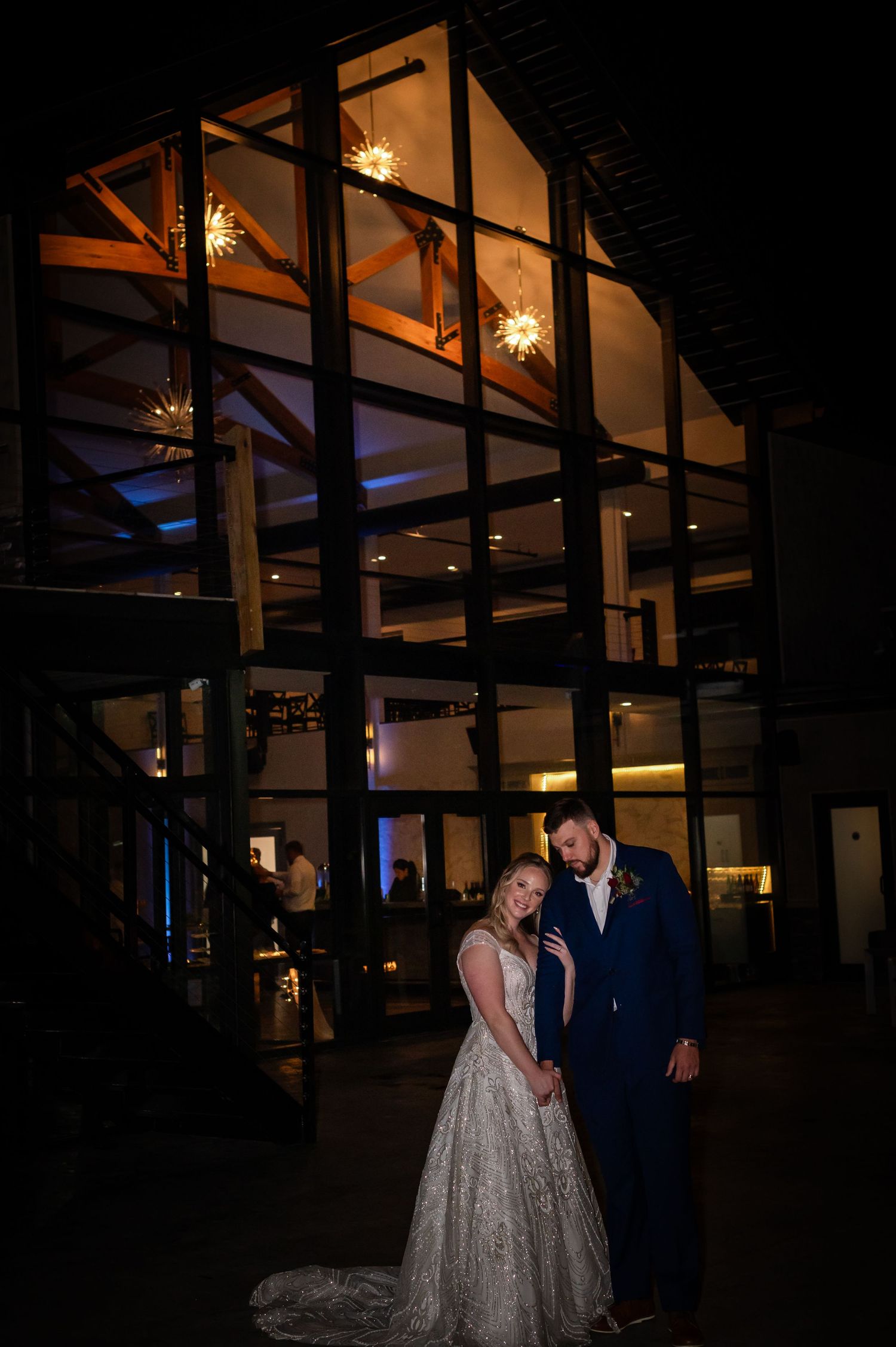 Evening wedding portrait with modern glass building facade lit with warm interior lights.