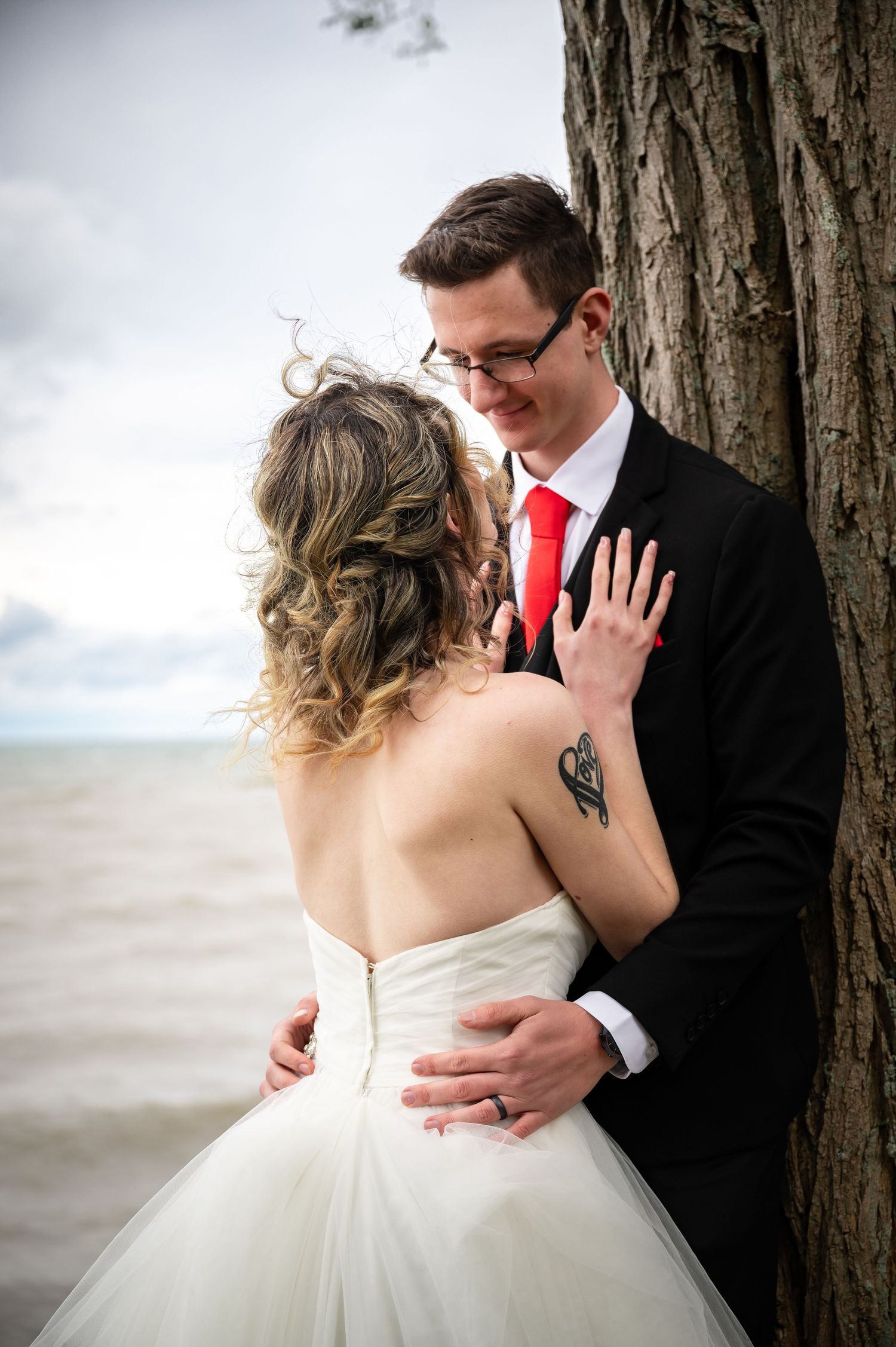 Intimate wedding portrait against a tree trunk by a stormy lake with dramatic cloudy skies.