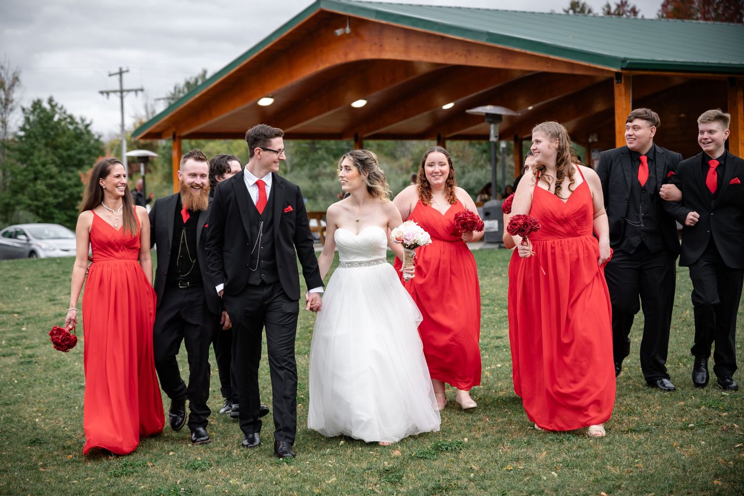 Wedding party laughing together outside pavilion wearing red dresses and black suits.
