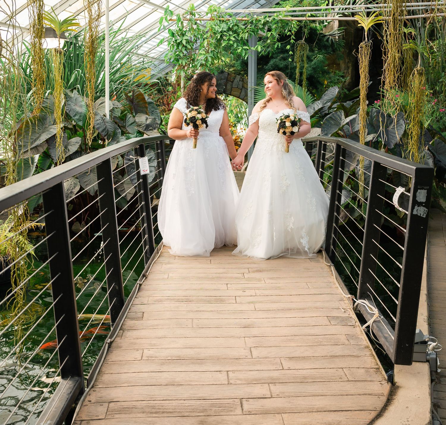 Two brides in white wedding dresses walk together on a wooden walkway surrounded by lush greenery in a greenhouse.