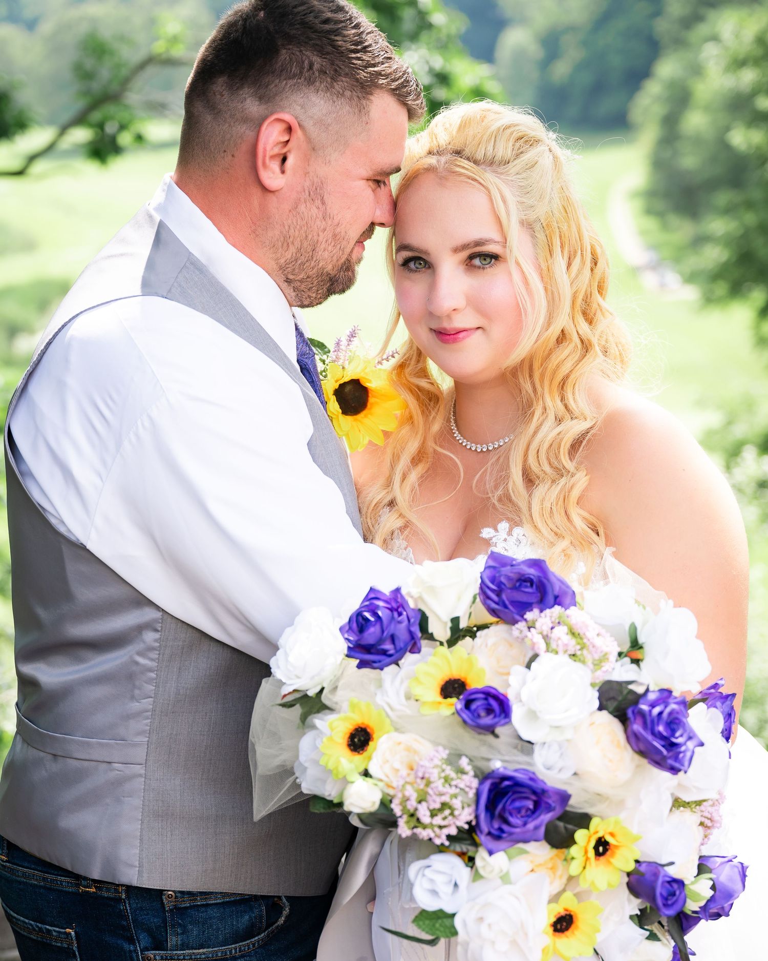 Close up of wedding couple with vibrant purple and yellow bouquet against green landscape.