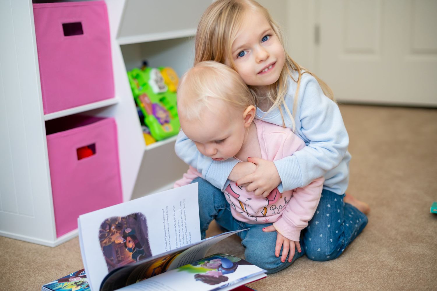 Two children looking at a book together near pink storage boxes.