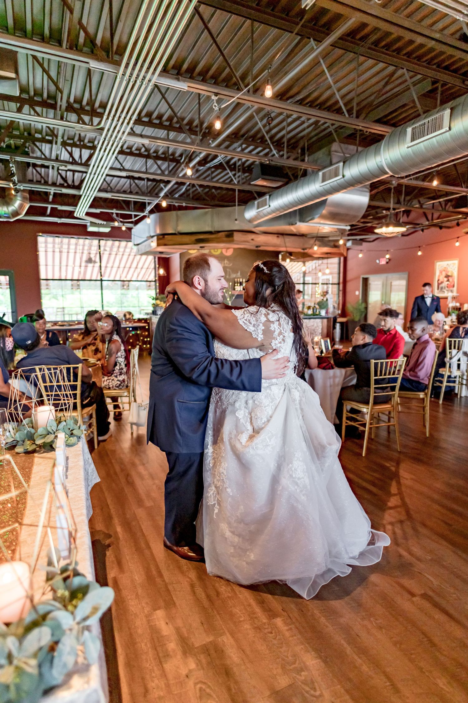 A romantic wedding dance on a hardwood floor with industrial ceiling details and elegant table settings.