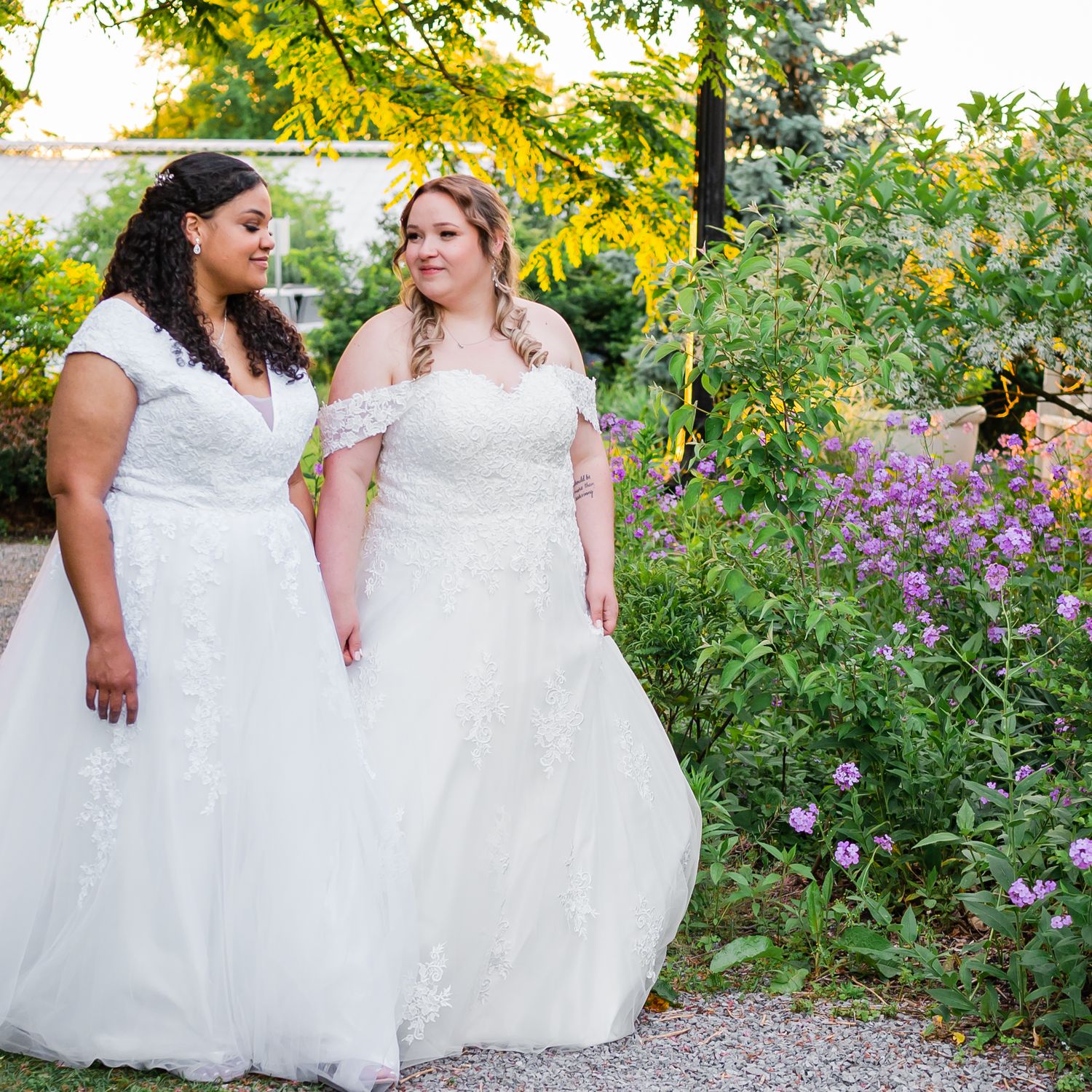 Two brides in elegant white wedding dresses stand together on a garden pathway with purple flowers.