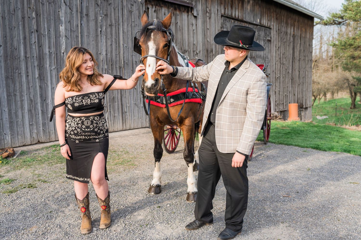 A couple poses with their horse outside a rustic barn wearing western-inspired outfits.