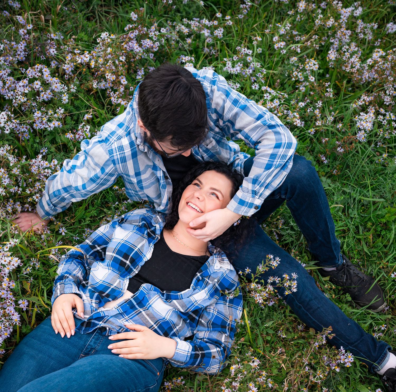 Two people in matching blue plaid shirts lay together among purple wildflowers in a meadow.