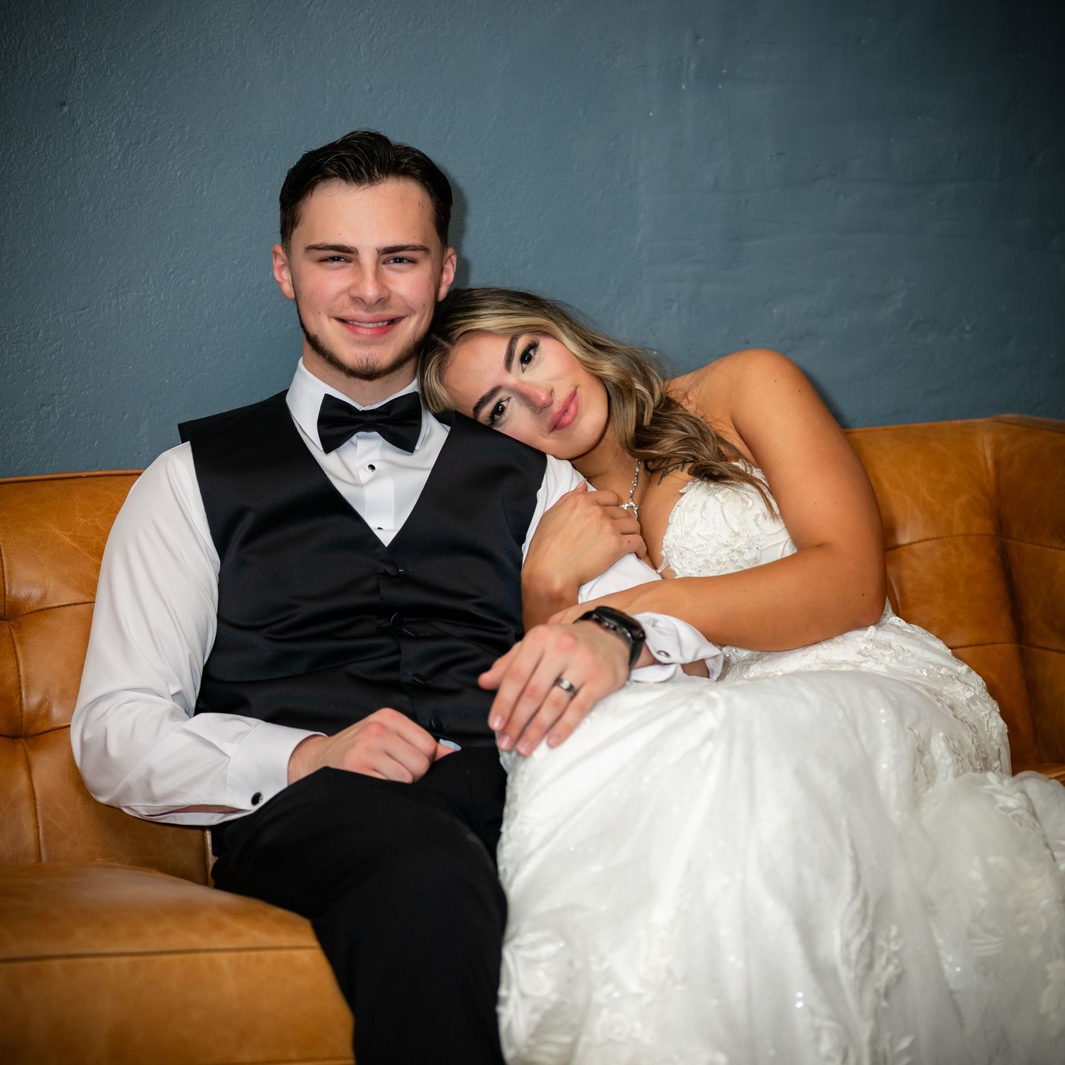 Newlywed couple relaxes together on a brown leather couch in formal wedding attire during their reception.