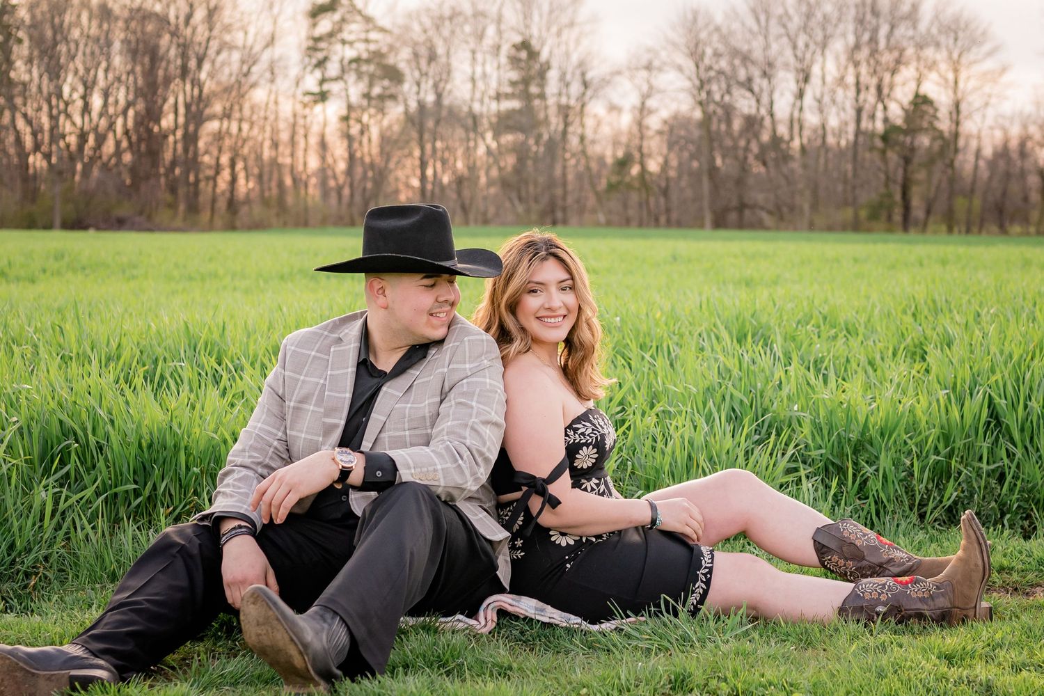 Two people sitting together on green grass in a field at sunset with rural scenery in the background.