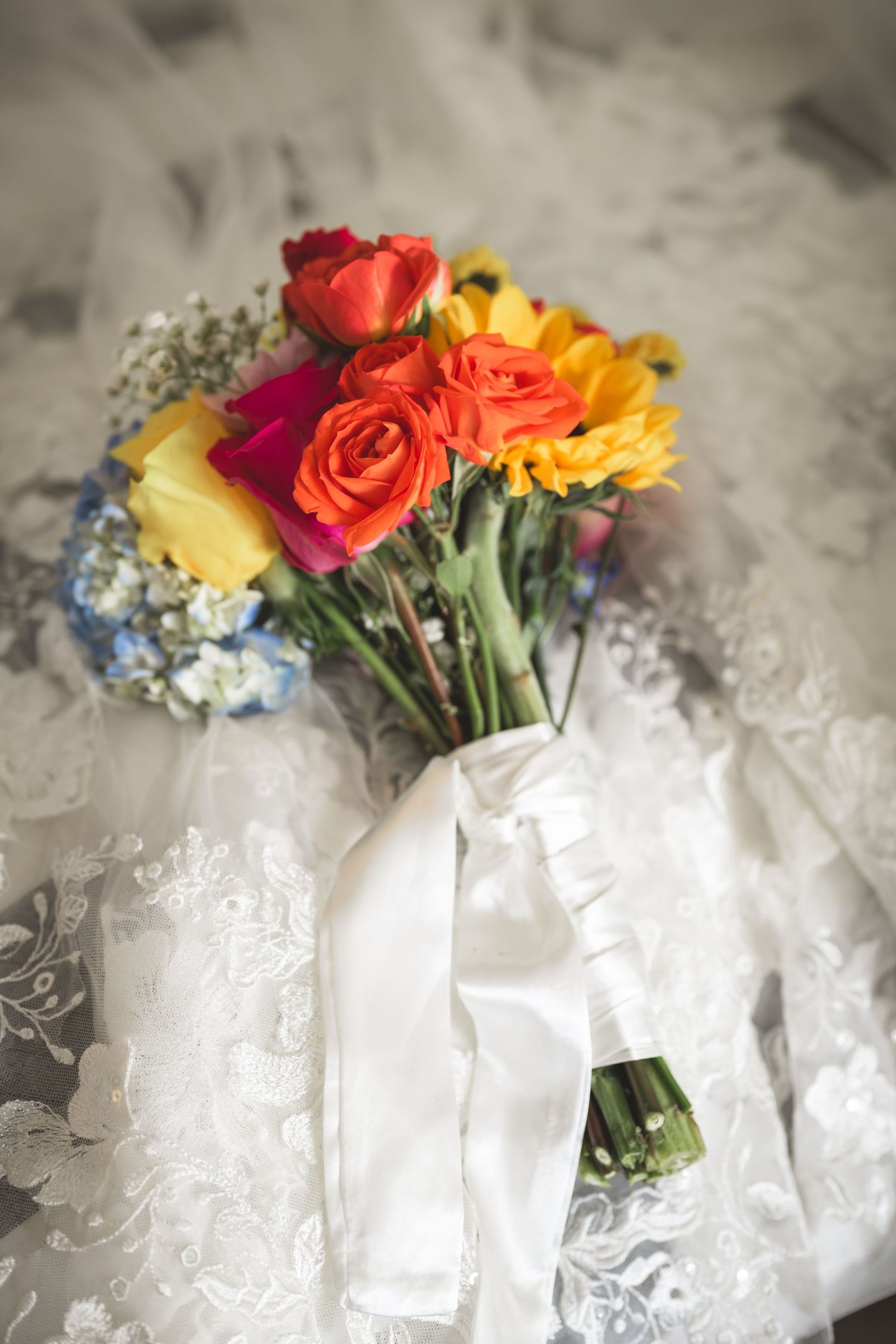 Colorful bridal bouquet with red, orange, and yellow roses tied with white ribbon on lace wedding dress.