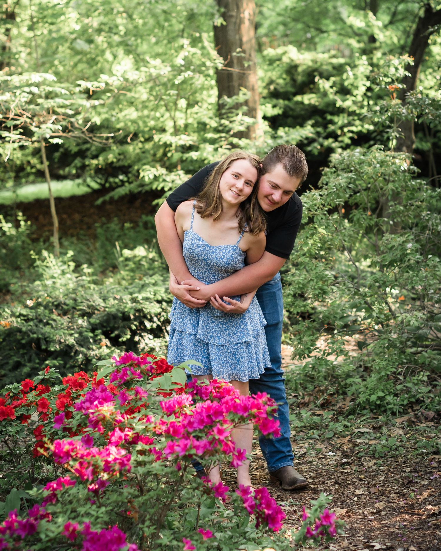 A couple embraces near vibrant pink flowers on a wooded garden path during a sunny summer day.