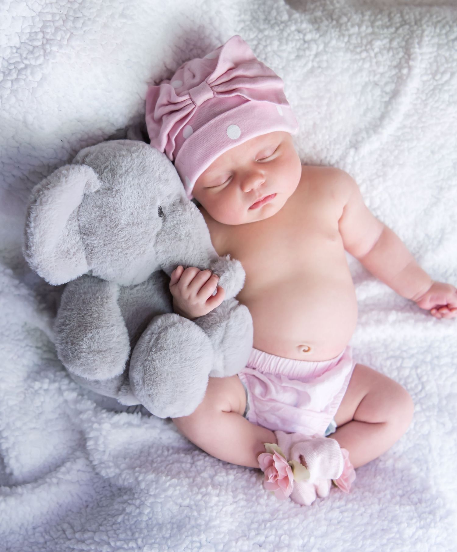 A sleeping baby in pink clothing lies on white bedding next to a plush gray elephant toy.