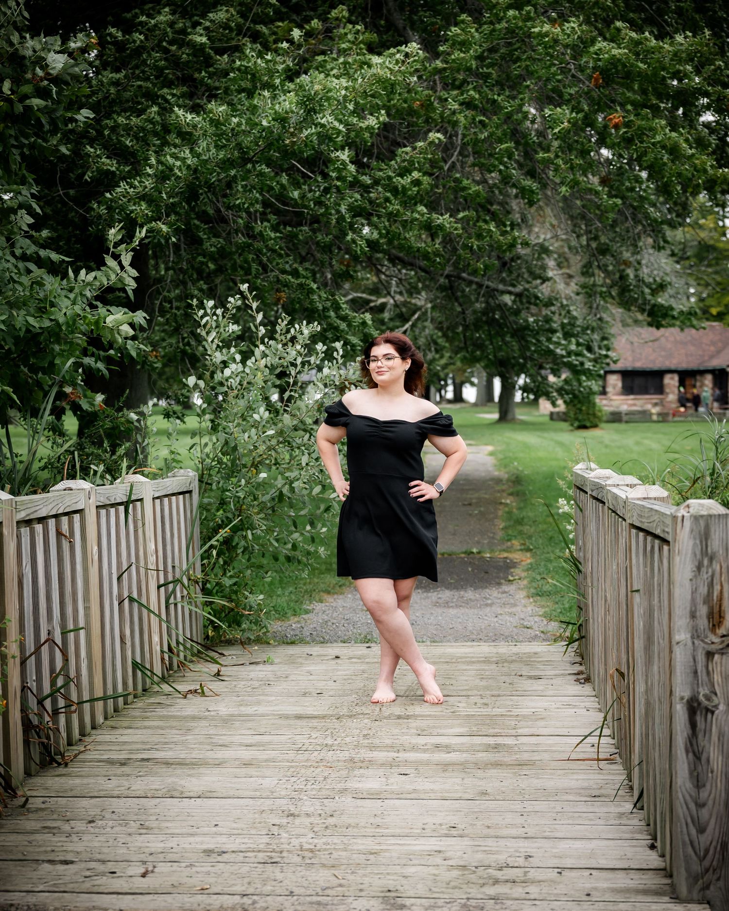 Person in black dress poses on wooden bridge surrounded by lush greenery.