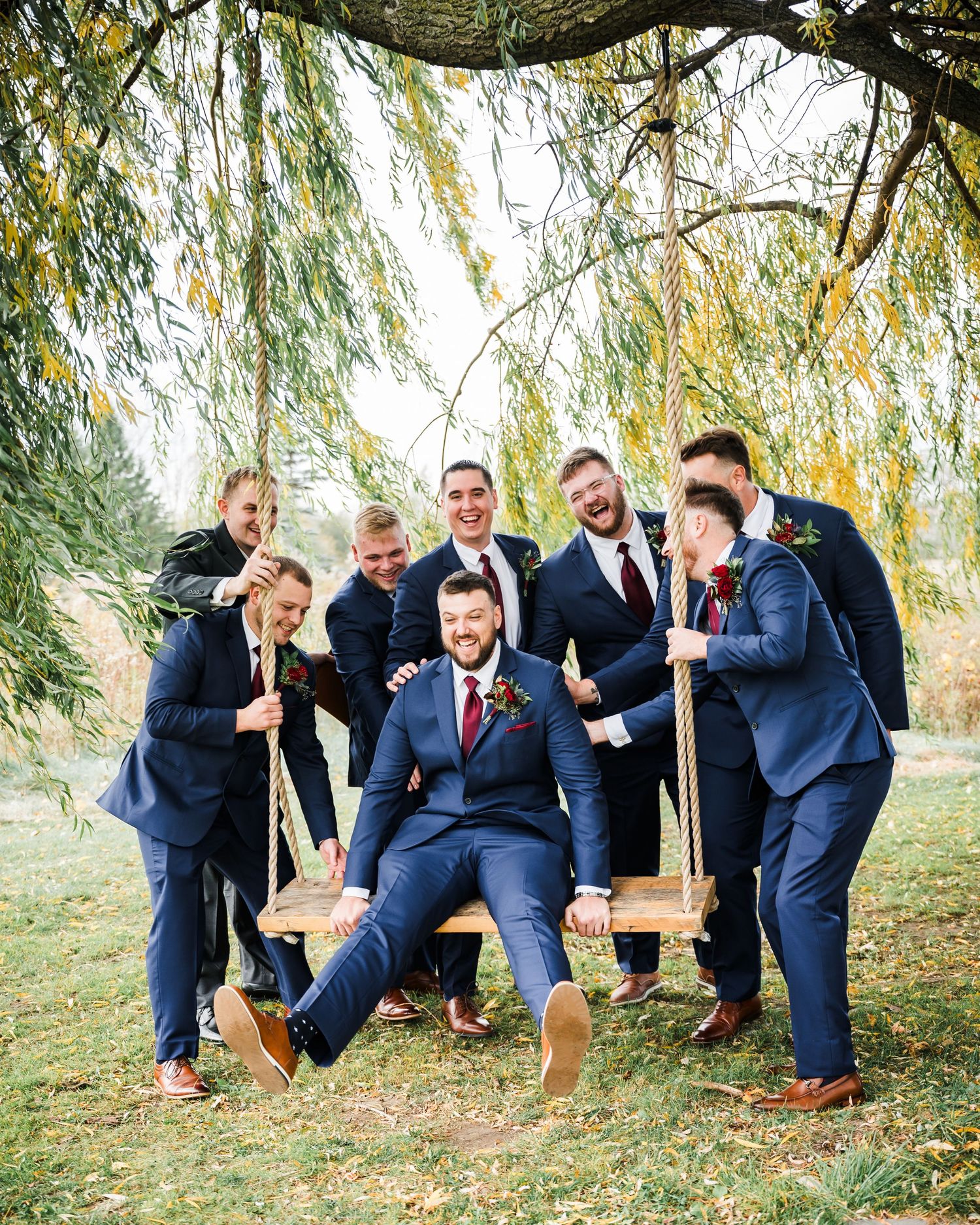 Groomsmen playfully pose with someone seated on a wooden swing under a willow tree.