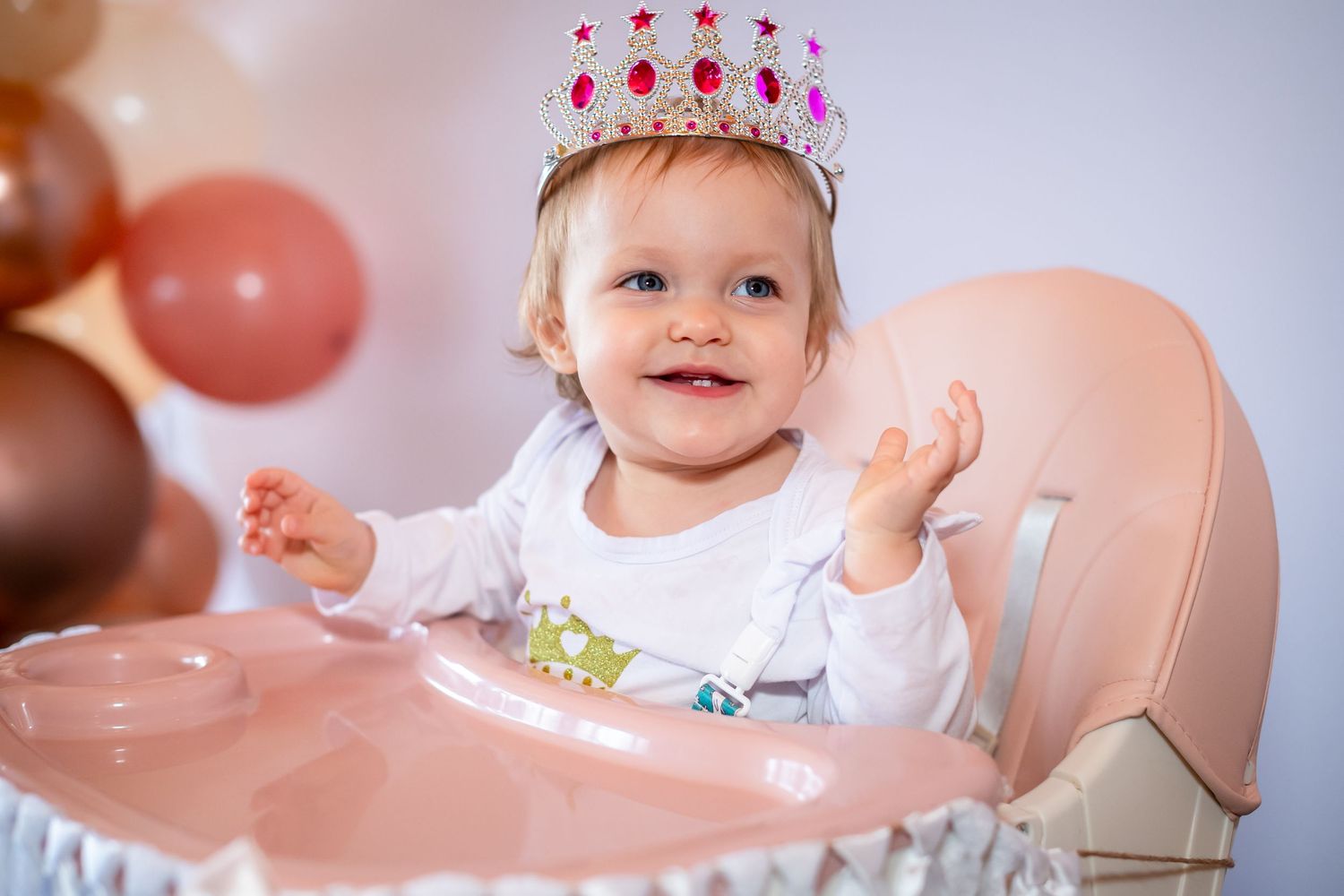 A baby wearing a crown celebrates their first birthday with pink balloons and decorations.