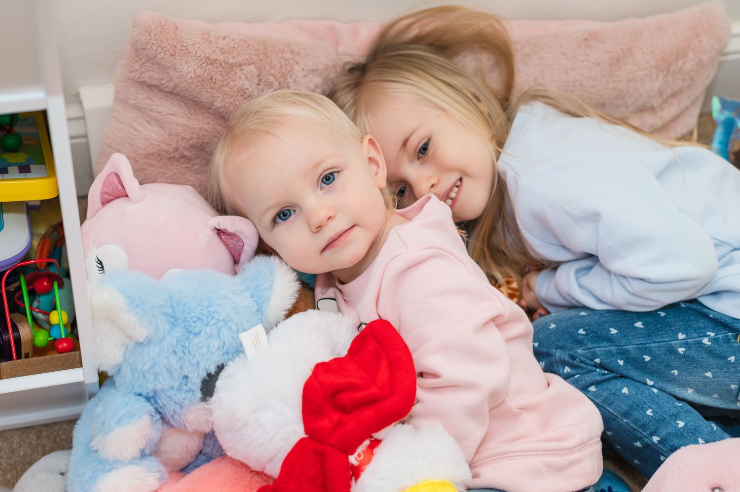 Two children cuddling with stuffed animals on pink blanket.