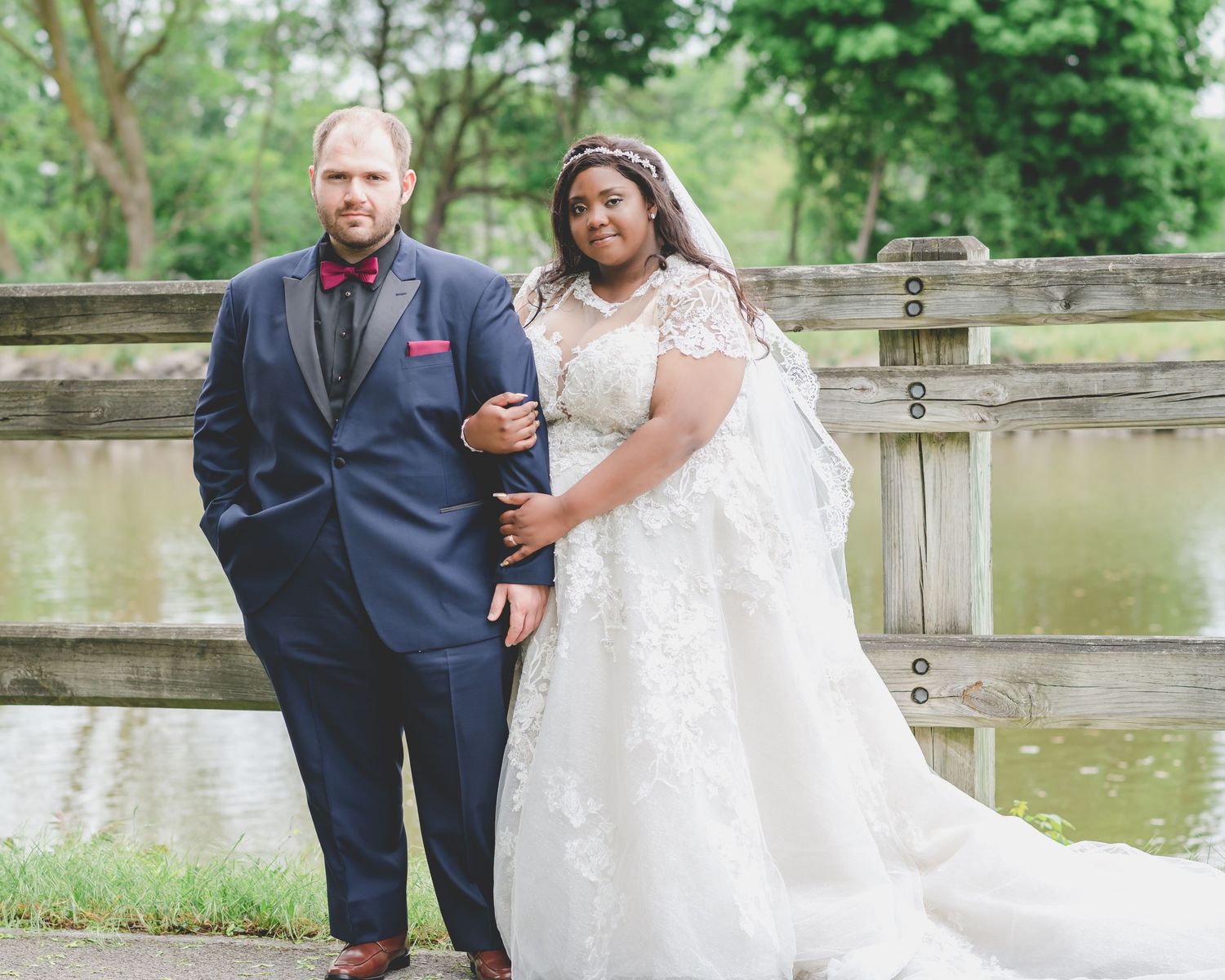 A wedding couple posing together in front of a wooden fence by a serene pond.
