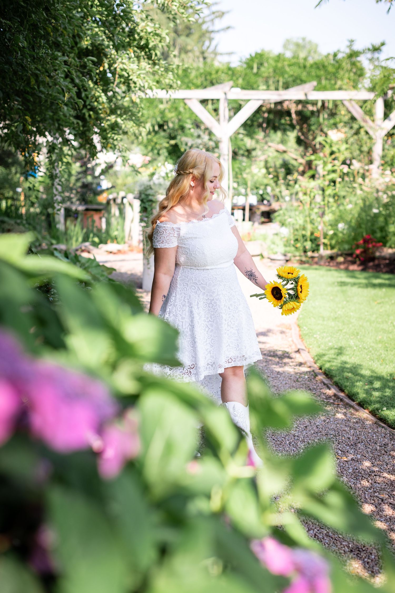 White dress-clad figure walks through garden path holding sunflower near wooden arbor.