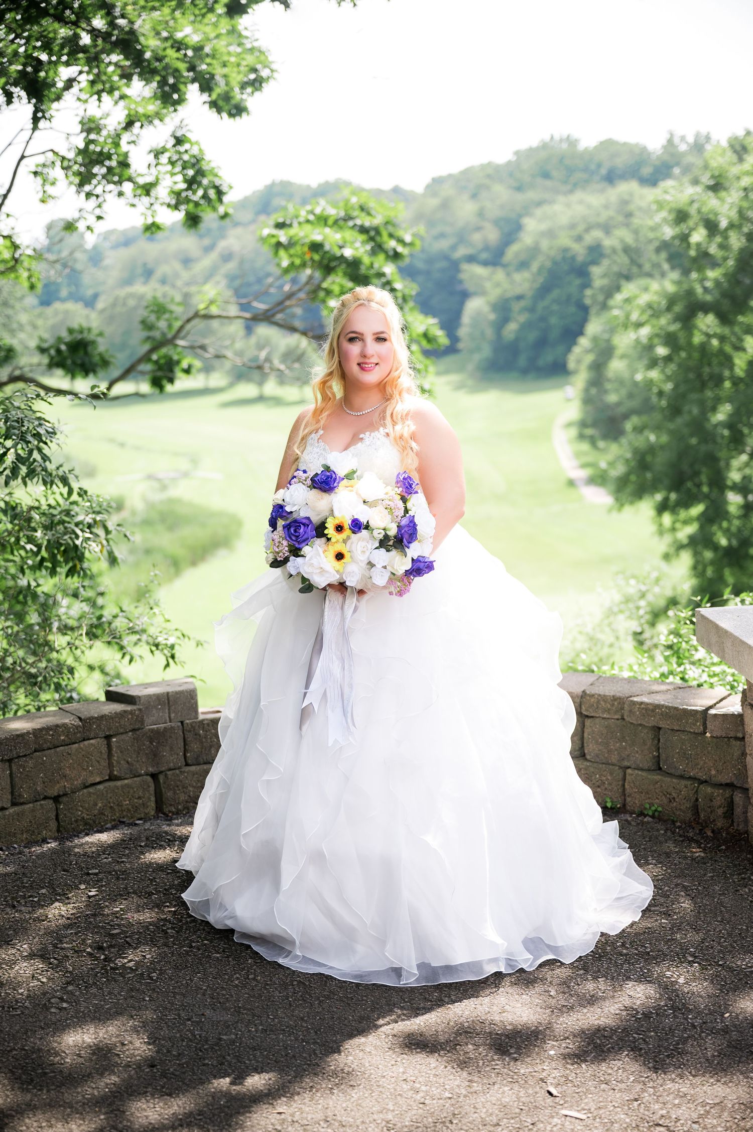A wedding portrait on a stone overlook showcasing a white ball gown and purple and yellow floral bouquet.