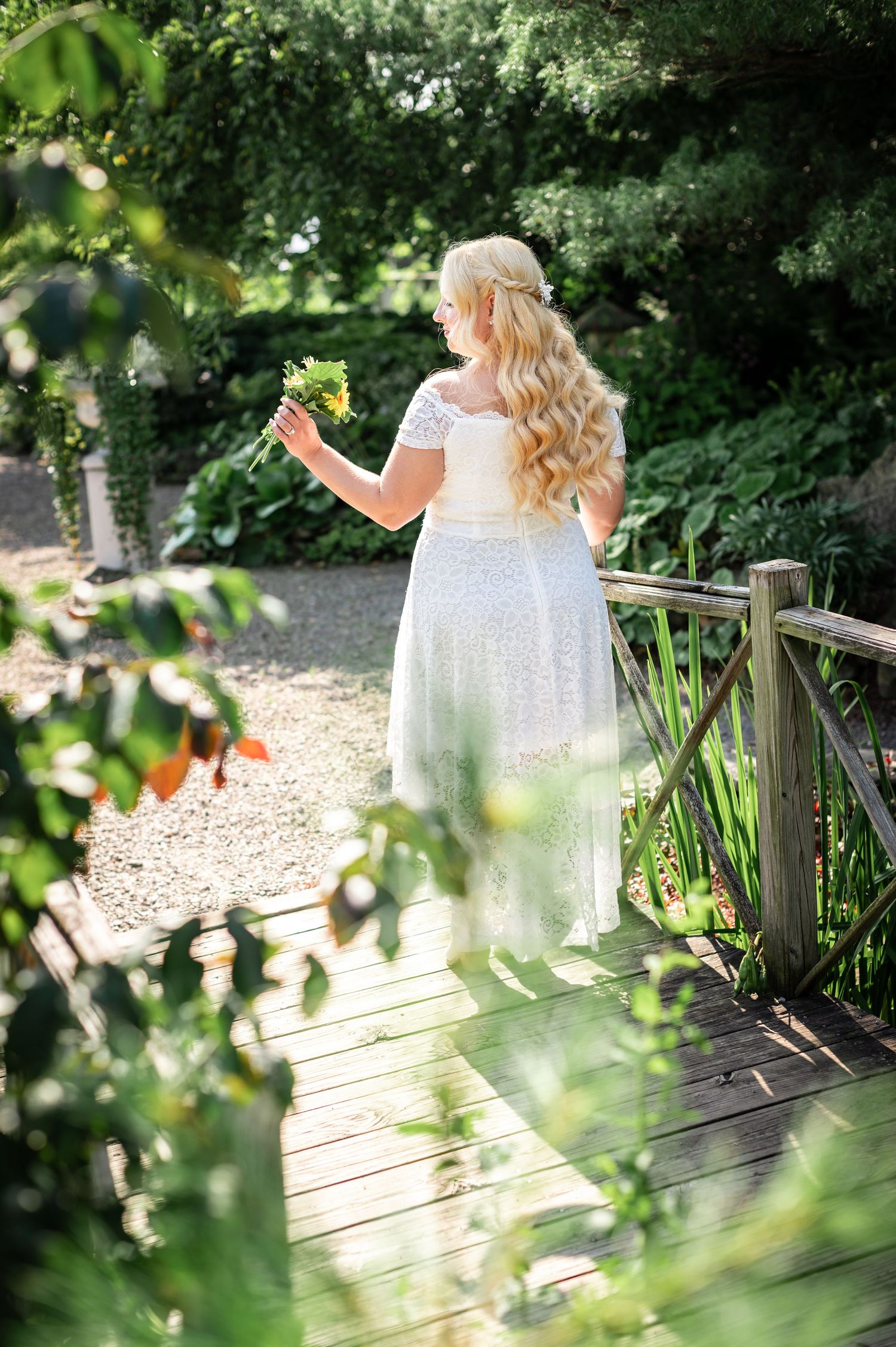 Person in flowing white dress stands on wooden bridge in lush garden setting.
