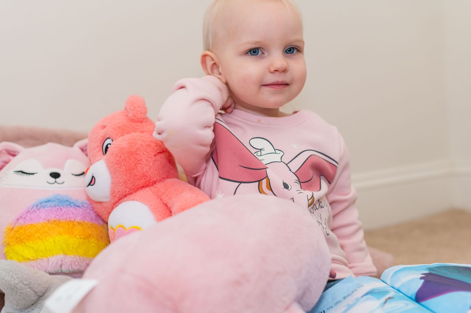 Child surrounded by colorful plush toys wearing pink sweatshirt with cartoon print.