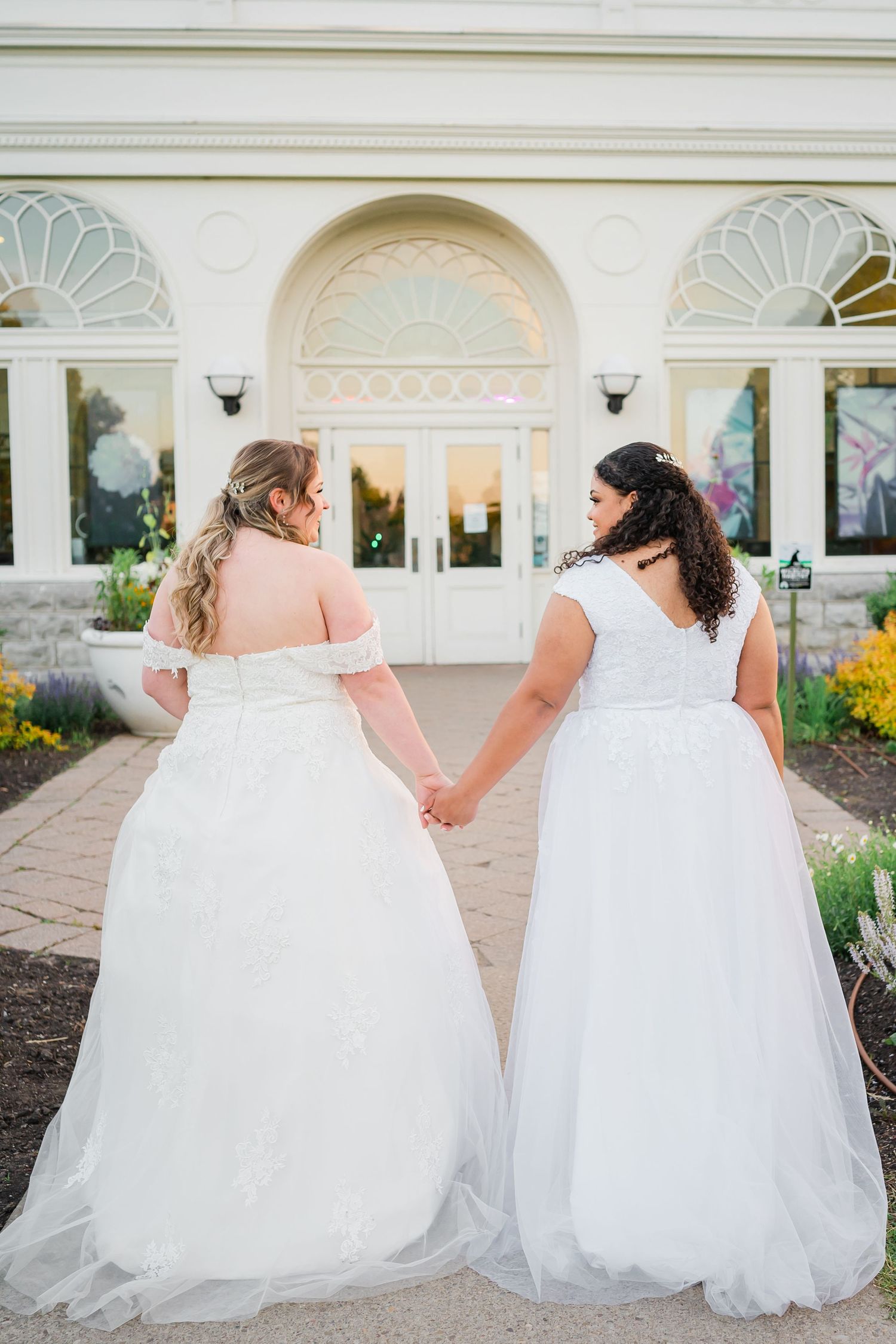 Two brides in white wedding dresses hold hands while standing in front of an elegant building entrance with floral displays.