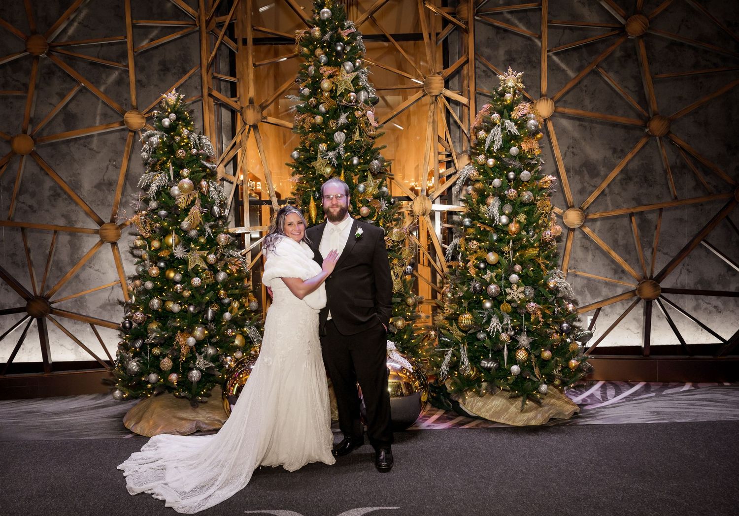 A wedding couple poses in front of three illuminated Christmas trees with elegant decorations.